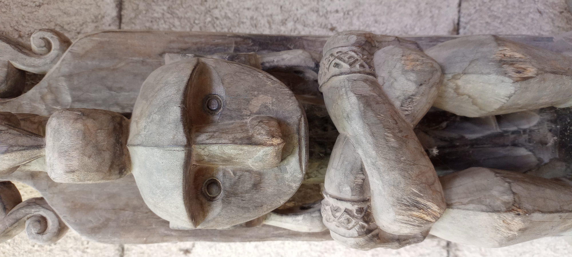 The image shows a close-up, eye-level shot of a weathered, wooden sculpture of a figure with a distinct, mask-like face. The sculpture is predominantly gray and brown, with visible wood grain and signs of age and wear. The figure has a large, rounded head with prominent, wide-set eyes that appear to be carved as shallow depressions. A long, angular nose dominates the center of the face, and a small, thin mouth is visible below. The figure is depicted with its arms crossed over its chest, resting on its lap. The arms are adorned with carved circular bands, possibly representing bracelets or armbands, featuring geometric patterns. The body of the figure is partially obscured by the crossed arms and the overall composition. The sculpture is mounted on a darker wooden back, which shows vertical lines indicating construction or carving. The background consists of a textured, light-colored surface, possibly a stone wall, which is out of focus. The lighting appears to be natural, casting soft shadows that highlight the textures of the wood. There is no discernible information in the image that would indicate its location in Gelsenkirchen, Germany, or any specific urban environment.