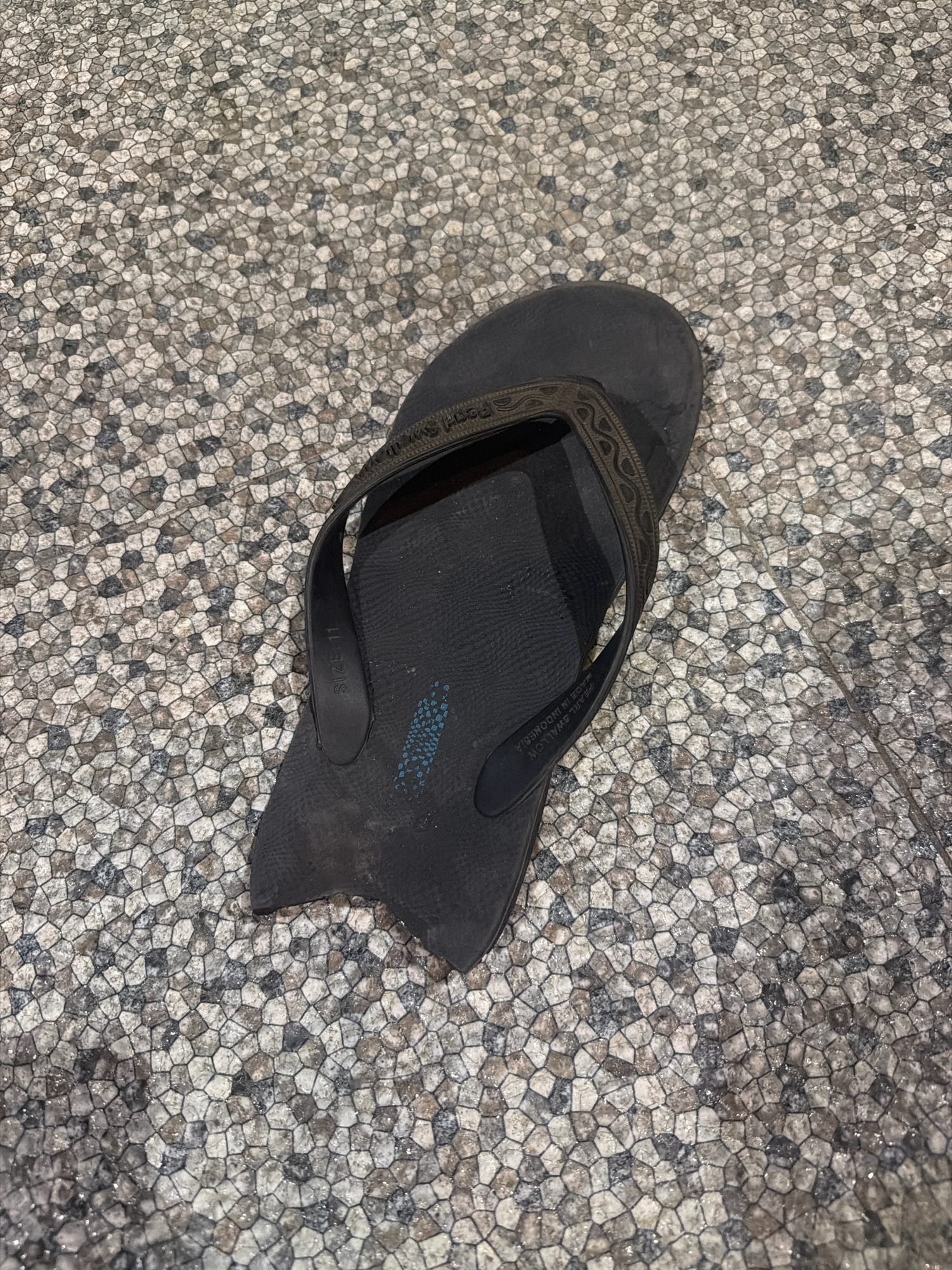 A black flip-flop lies on a textured stone floor. The flip-flop is oriented diagonally, with the sole facing upwards. The sole has some blue markings and appears worn. The strap of the flip-flop is also black and has a textured pattern. The stone floor is composed of small, irregularly shaped stones in various shades of grey, white, and brown, creating a mosaic-like pattern. The overall lighting suggests an indoor or shaded outdoor environment. This scene is likely located in Balikpapan, Indonesia, given the provided context, possibly on a patio, walkway, or entrance area.