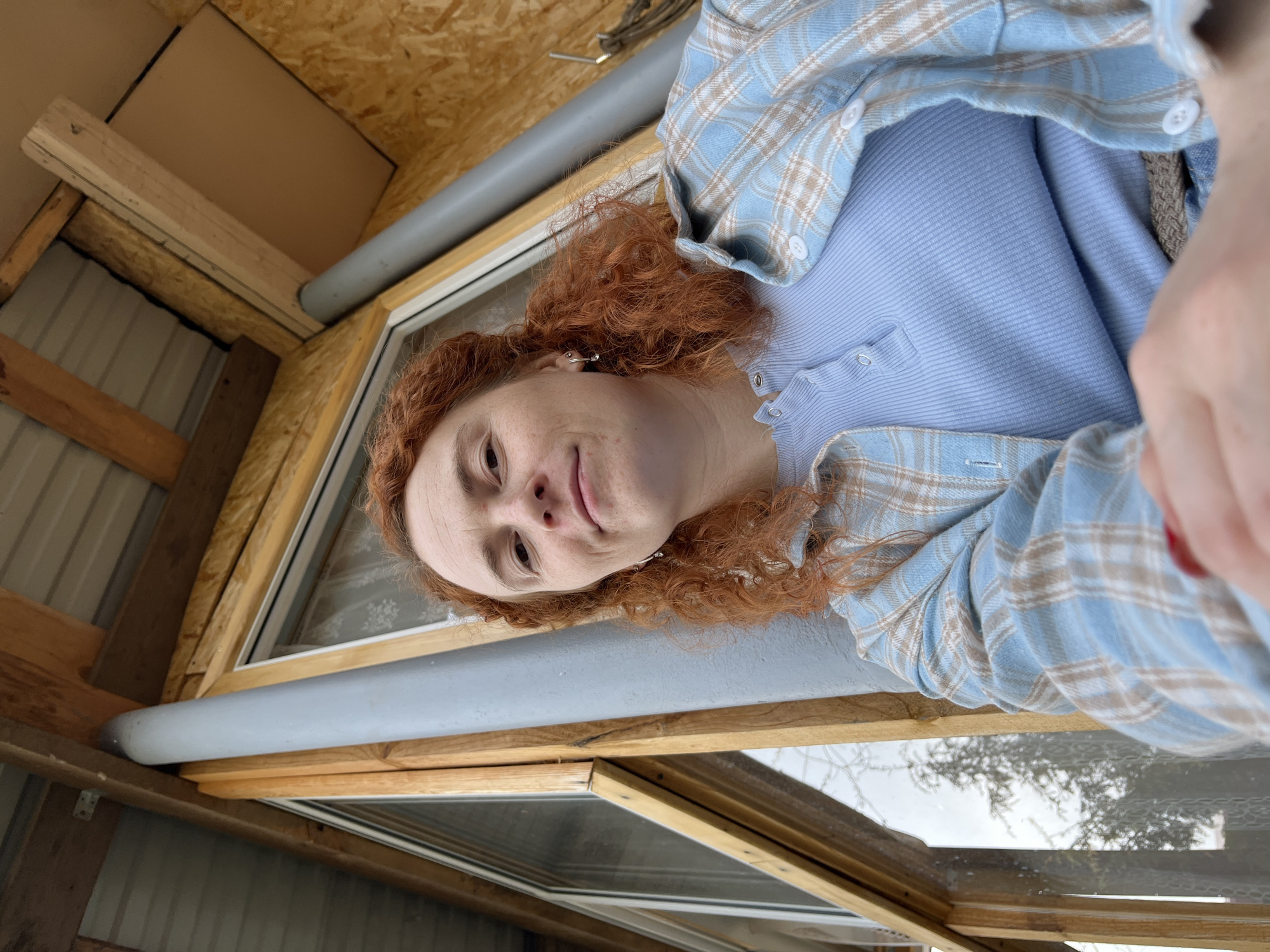 A medium shot presents a person with fair skin and reddish-brown curly hair, wearing a light blue button-down shirt over a matching blue top. The person is indoors, positioned in what appears to be an unfinished room or structure, potentially in the city of Chaikovsky, Russia. The room's construction features exposed wooden beams and plywood panels. A window with a wooden frame and visible sealant is in the immediate foreground, suggesting recent construction or renovation work. The background through the window shows bare trees against a cloudy sky, indicating a possible winter or early spring setting. A gray cylindrical object runs along the window frame. The person is looking slightly down at the camera with a faint smile.