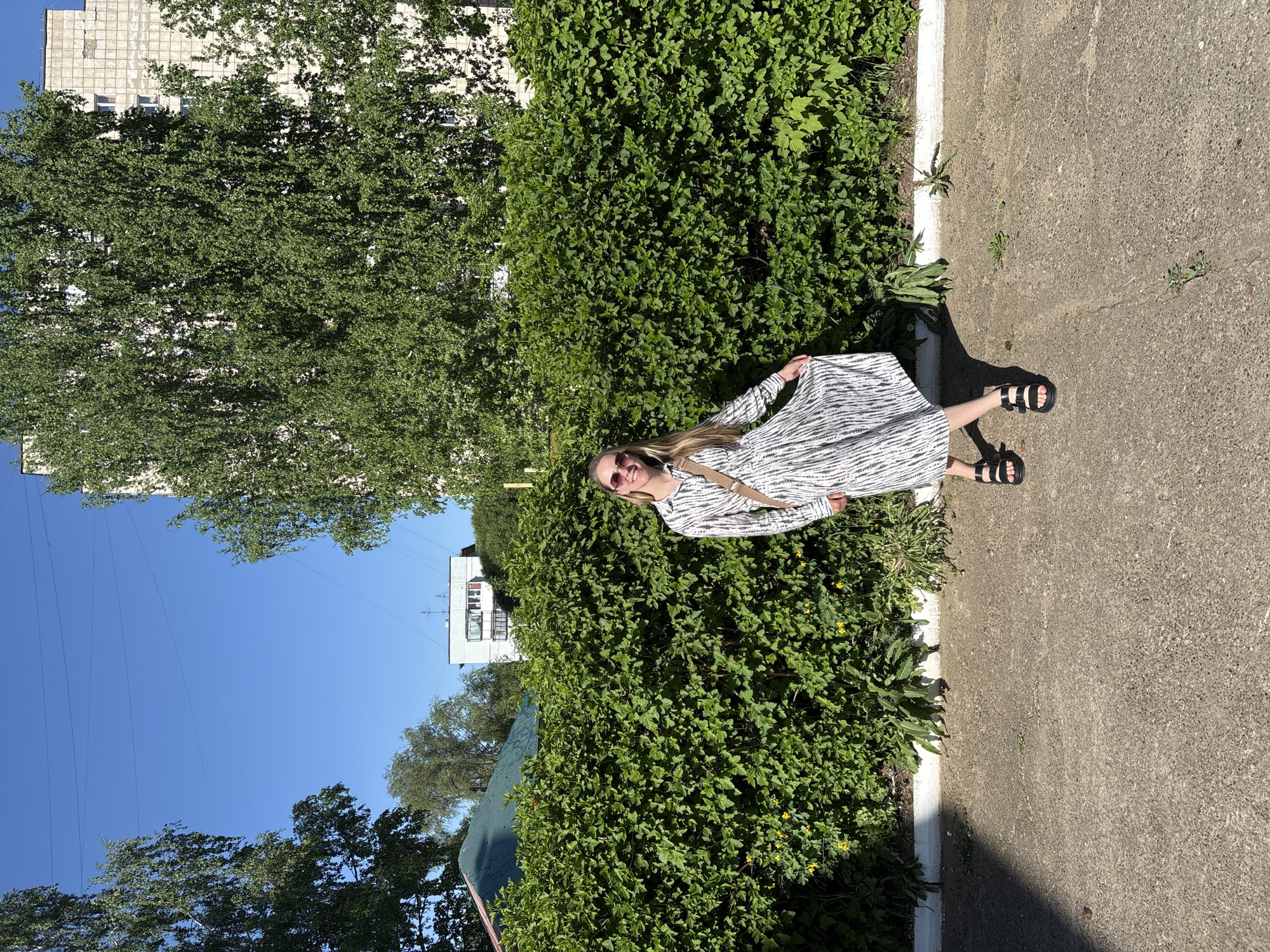 A young woman stands in front of a tall, dense green hedge, smiling. She wears a striped, light-colored dress, sandals, and sunglasses. She is fair-skinned with long blonde hair. She holds the dress with both hands. A building is visible behind the tree. The building appears to have a balcony with potted plants. Above, powerlines are visible against a clear blue sky. A paved pathway runs along the edge of the hedge. The location appears to be outdoors on a sunny day in an urban area. Without more precise information, the exact location in the city of Chaikovsky cannot be determined.
