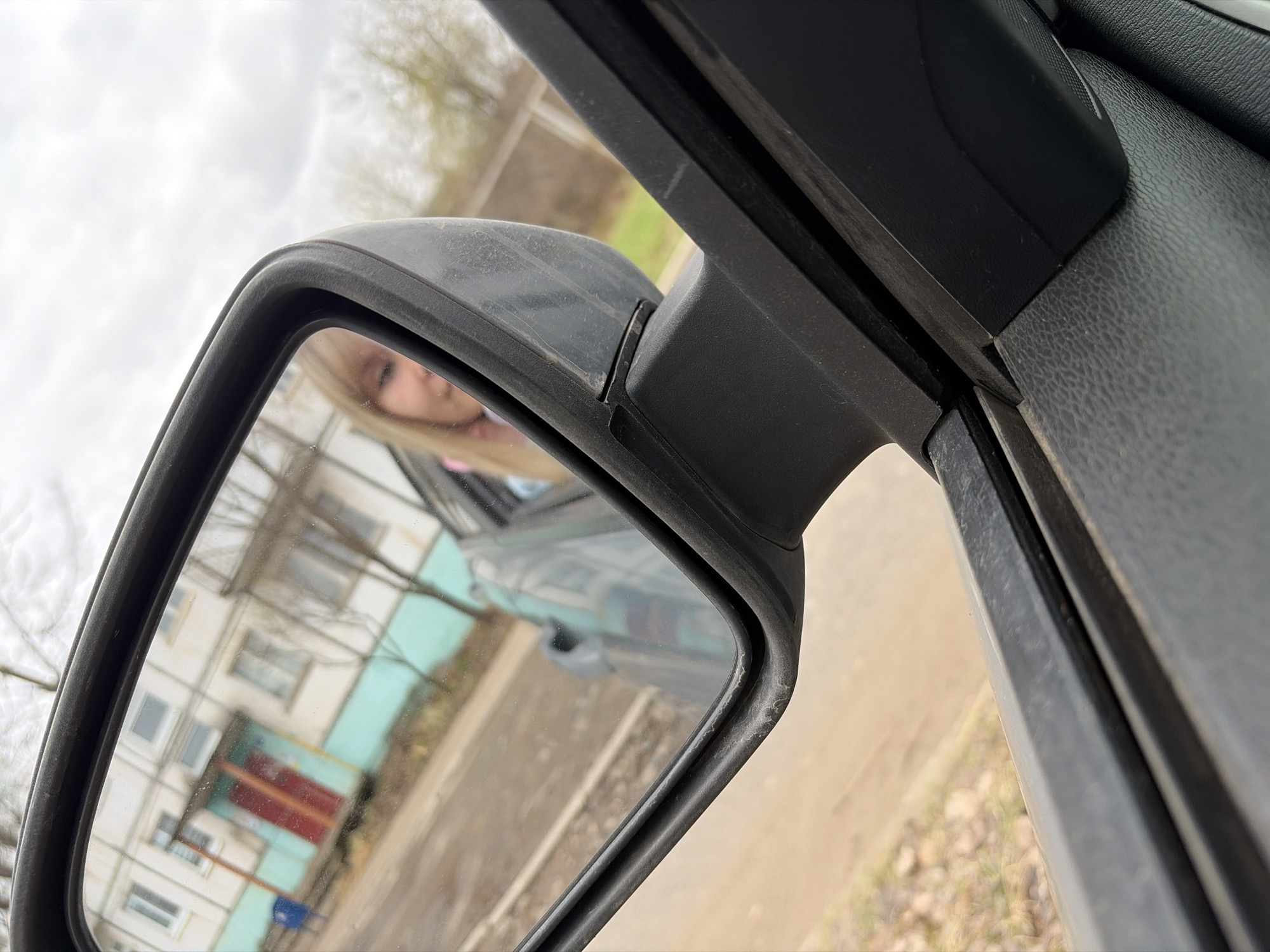 The image shows the side mirror of a car, reflecting a building and a woman with blonde hair. The building is light-colored with a red garage door. The sky is overcast. The immediate foreground contains the car door's interior panel. The location appears to be a residential area in an unspecified city.