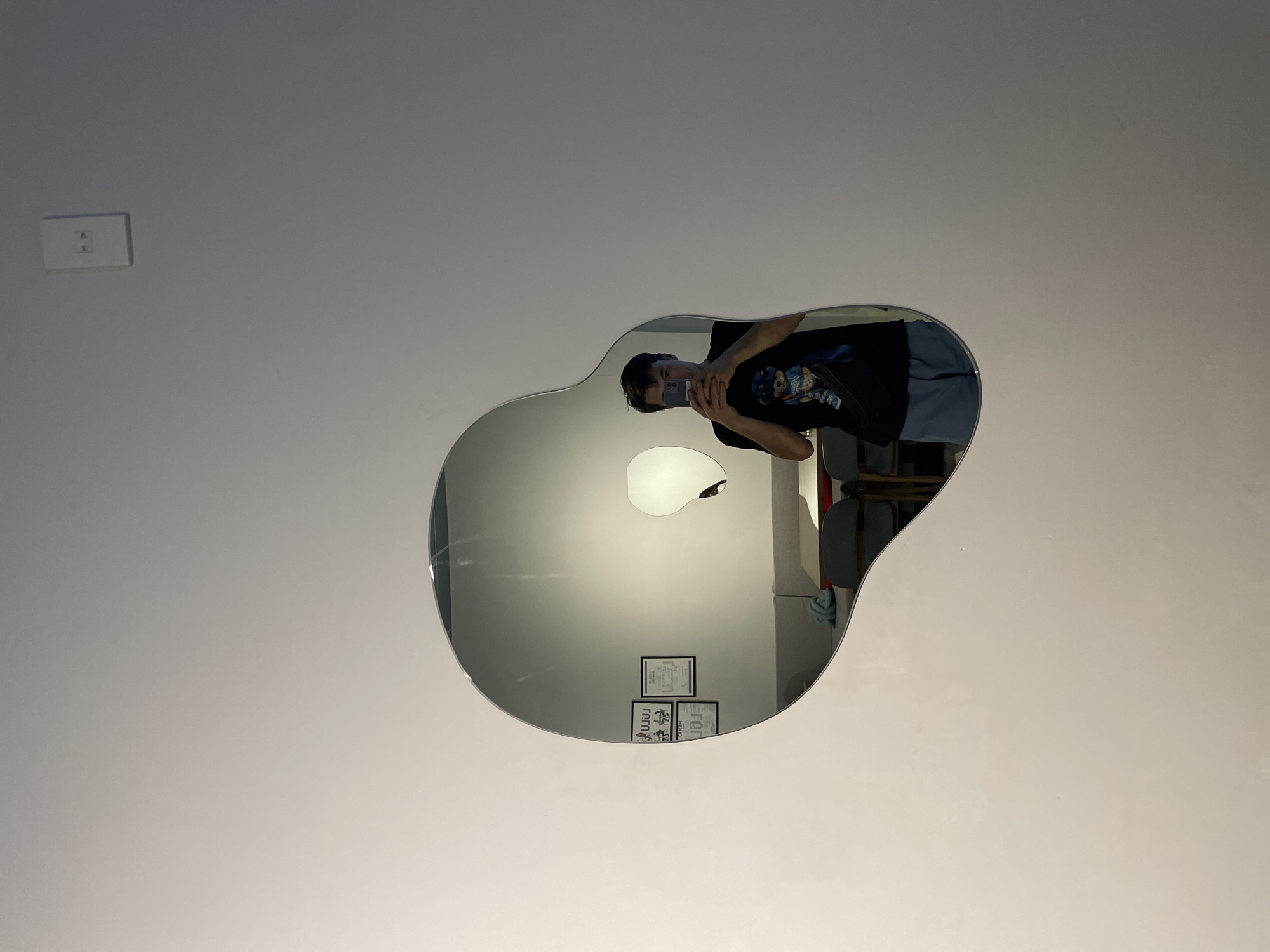 The image displays an indoor scene featuring a person taking a mirror selfie. An irregularly shaped, reflective mirror, resembling an organic or blob-like form, is mounted on a plain, light-colored wall.
Reflected in the mirror is a person, appearing to be male, holding a smartphone to capture their reflection. They are wearing a dark t-shirt with a blue and white graphic design of what looks like a cartoon character. Their face is partially obscured by the phone. Above and slightly behind the reflected person, a bright, circular light source (likely a ceiling light) is visible within the mirror's reflection. Below this light, on what appears to be a wall behind the camera's position, are four small framed items or posters. One clearly shows the text "HERO" and another "UJ JU" or "ruru".
To the left of the mirror, an off-white electrical wall socket with two horizontal slots is visible on the wall. Hints of other furniture or objects, possibly a chair and a light blue cloth, are visible in the right side of the mirror's reflection. The overall setting is simple, suggesting a room in a home or apartment. The image captures a casual moment of self-portraiture. This scene is located in Bắc Nha Trang, Vietnam.