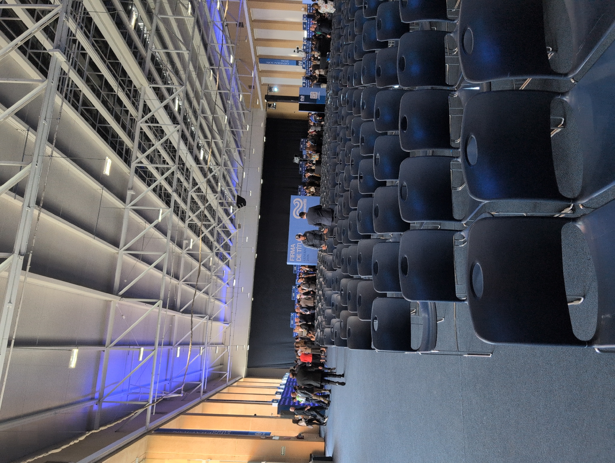 The image depicts an indoor conference or event space in Mexico City, Mexico. Rows of unoccupied black chairs face a stage area where people are seated and standing. Banners with text and logos are visible on the stage. The building's structural steel ceiling is exposed. Some of the individuals are wearing business attire.