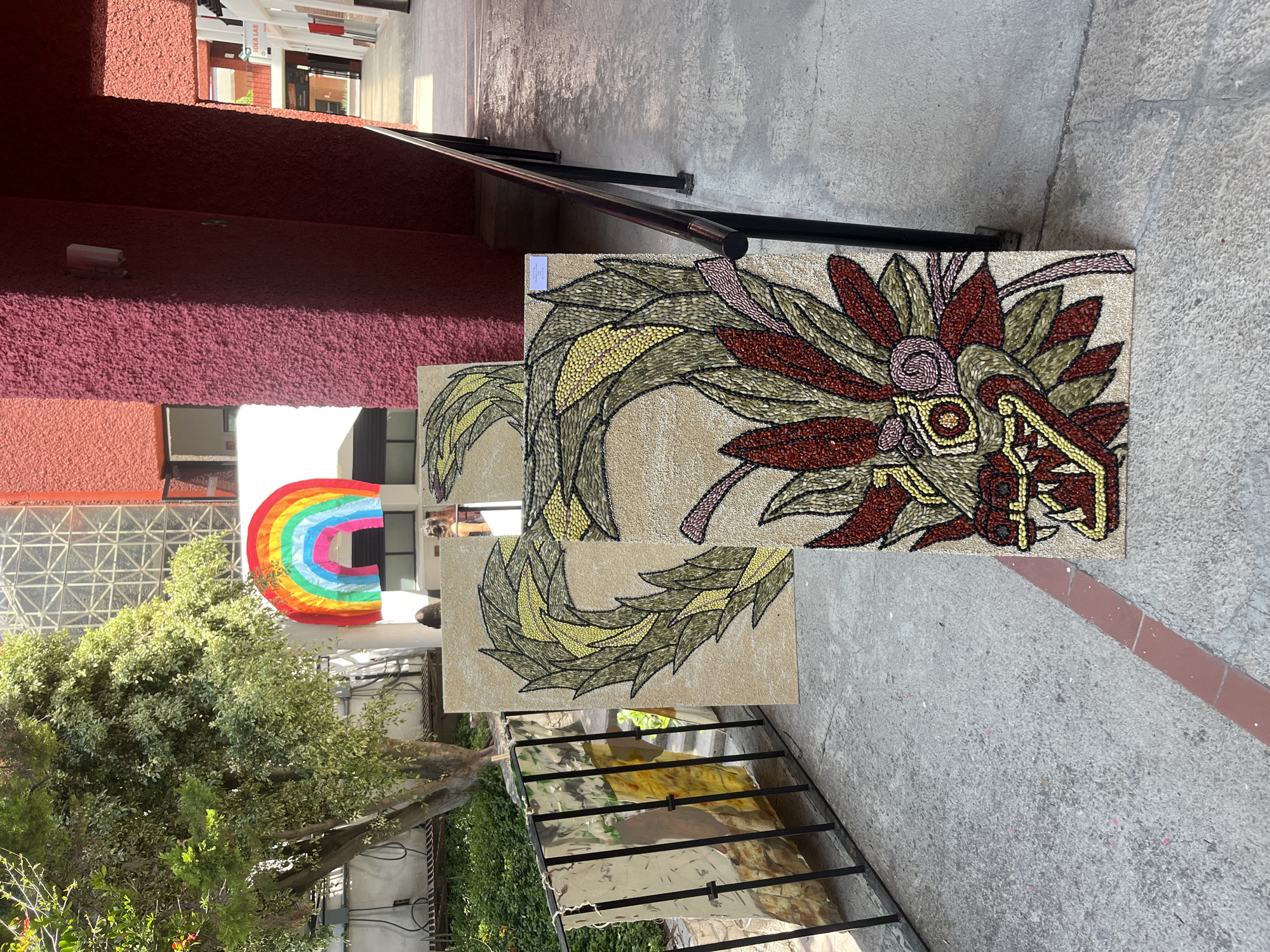 Outside a red-walled building in San Andrés Cholula, Mexico, a walkway displays two rectangular artworks propped against the wall. The artworks, seemingly mosaics, depict a serpent with detailed, ornate head and tail. The artwork is composed of small pieces, likely beans or seeds, in various colors including red, yellow, brown, and green. To the left of the mosaic, through greenery, a white building features a rainbow decoration. A metal handrail partially obscures the lower left of the scene, overlooking a lower level. The walkway is concrete, showing signs of weathering. A small sign is attached to the top piece.