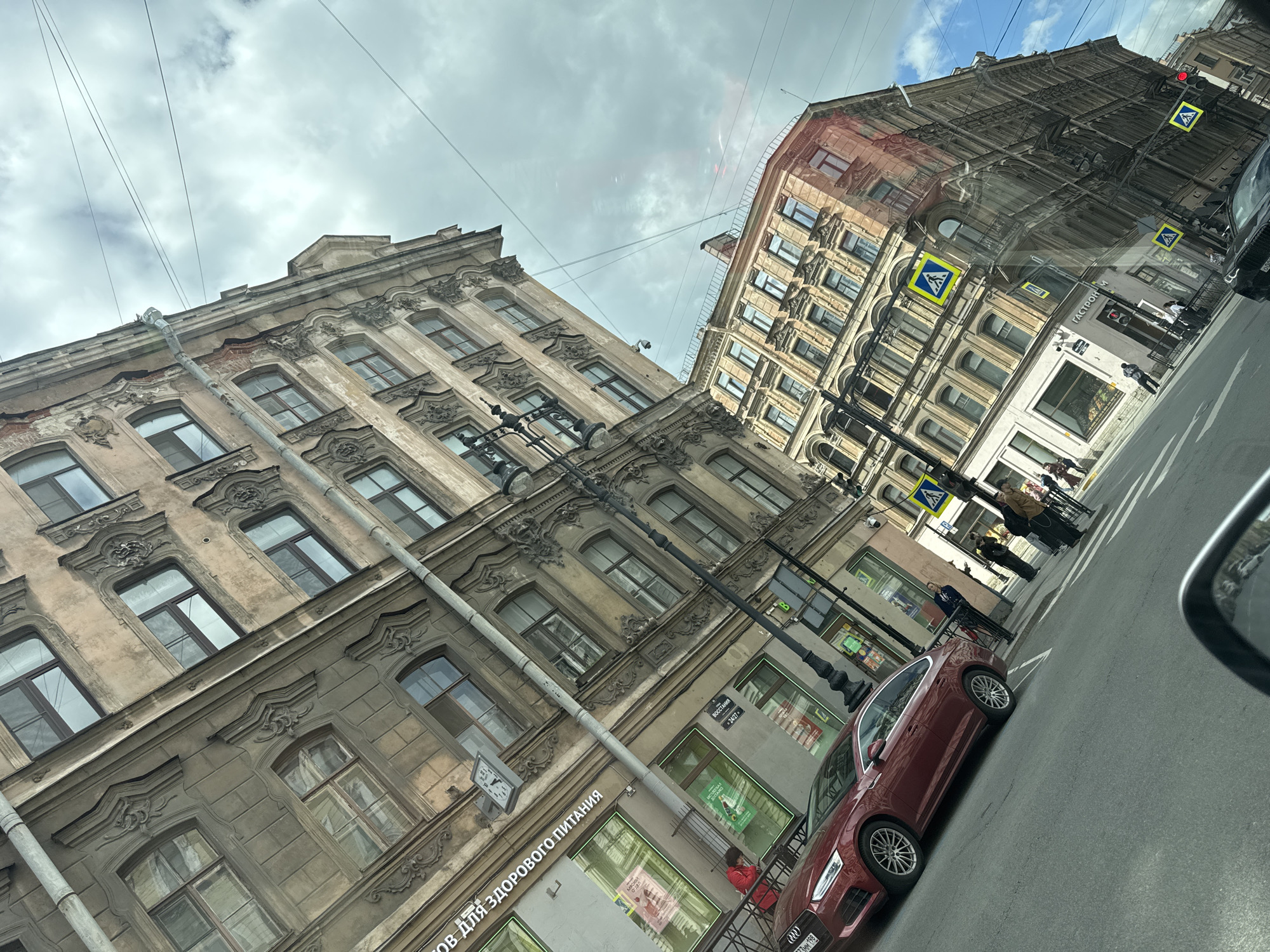 The image captures a street view in Saint Petersburg, Russia. Two buildings with classical architectural details dominate the scene. The building on the left has ornate window frames and a weathered facade. A clock and a sign in Russian, “Товаров для здорового питания” are visible on the building's side. The building on the right displays a similar architectural style. Pedestrian crossing signs indicate a crosswalk in front of the building.
A red car is parked on the street. A person in a red jacket sits on a fence near the car. People are walking on the sidewalk, including individuals with luggage. Utility poles with wires crisscross above the buildings. The sky is partially cloudy with patches of blue. A car is visible in the reflection of the car window.