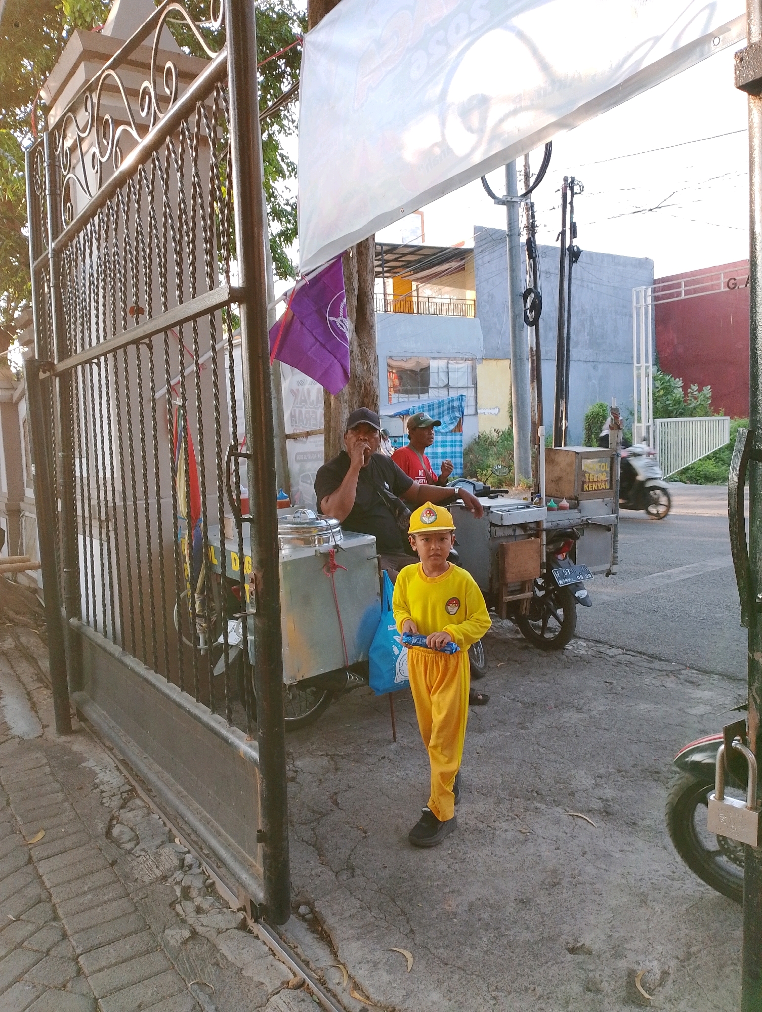 A young boy wearing a yellow jumpsuit and cap stands on a cracked concrete sidewalk in front of a food cart. He holds a blue toy in his hands. The food cart, which appears to be a mobile vendor, has metal containers and a wooden top. Two men are visible behind the cart, one wearing a black shirt and cap, and the other wearing a red shirt and green cap. A purple flag hangs from a pole behind them. In the background, a motorcycle is seen driving away, and buildings are visible. The scene suggests a street-side food vendor operation in a city environment.