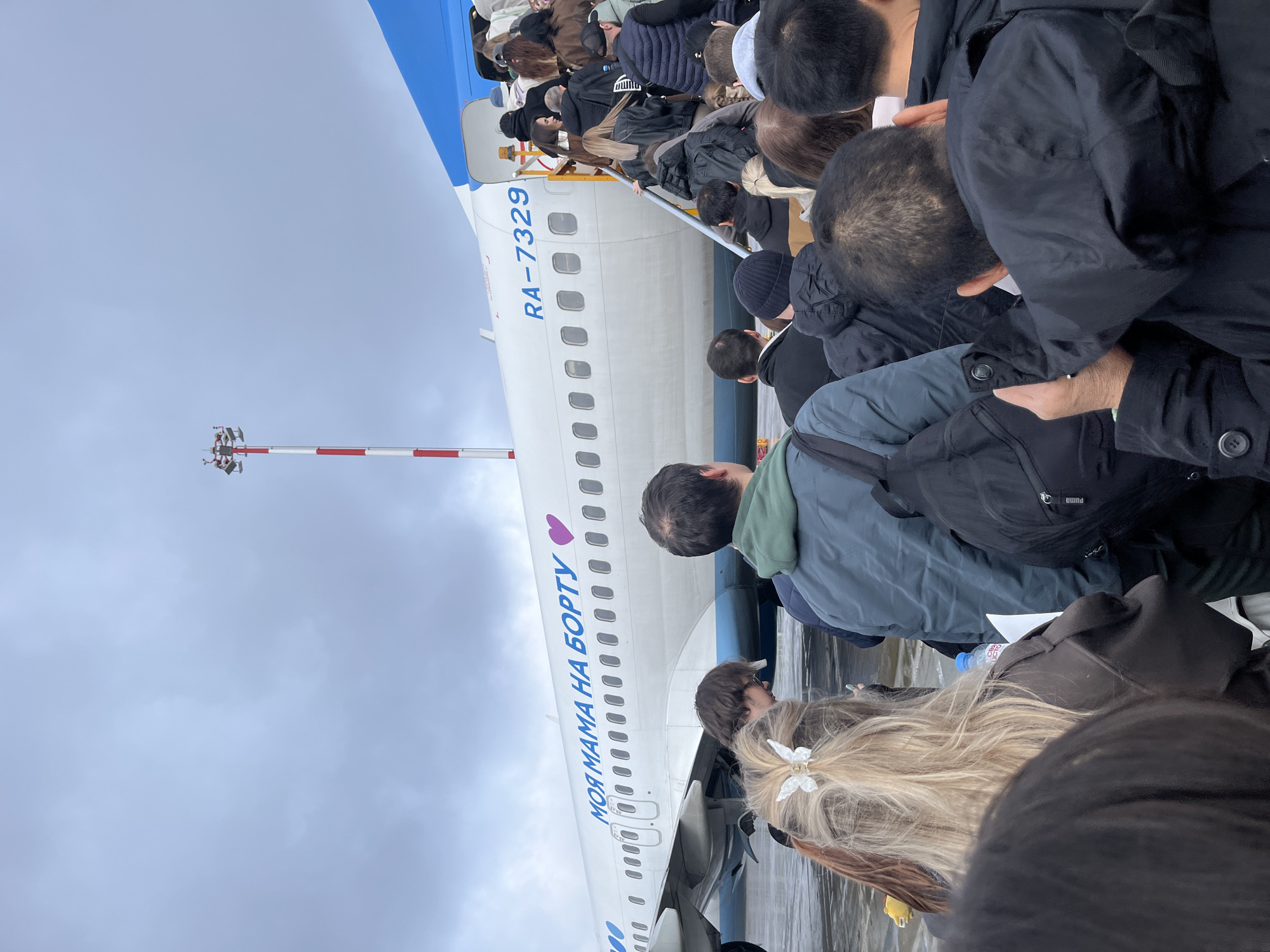 Passengers are boarding a white airplane with a blue stripe, registration number RA-7329. The plane has "МОЯ МАМА НА БОРТУ" written on it in blue, with a purple heart replacing the "O" in "МОЯ". A tower with red and white stripes is visible in the background. The location is Khimki, Russia.