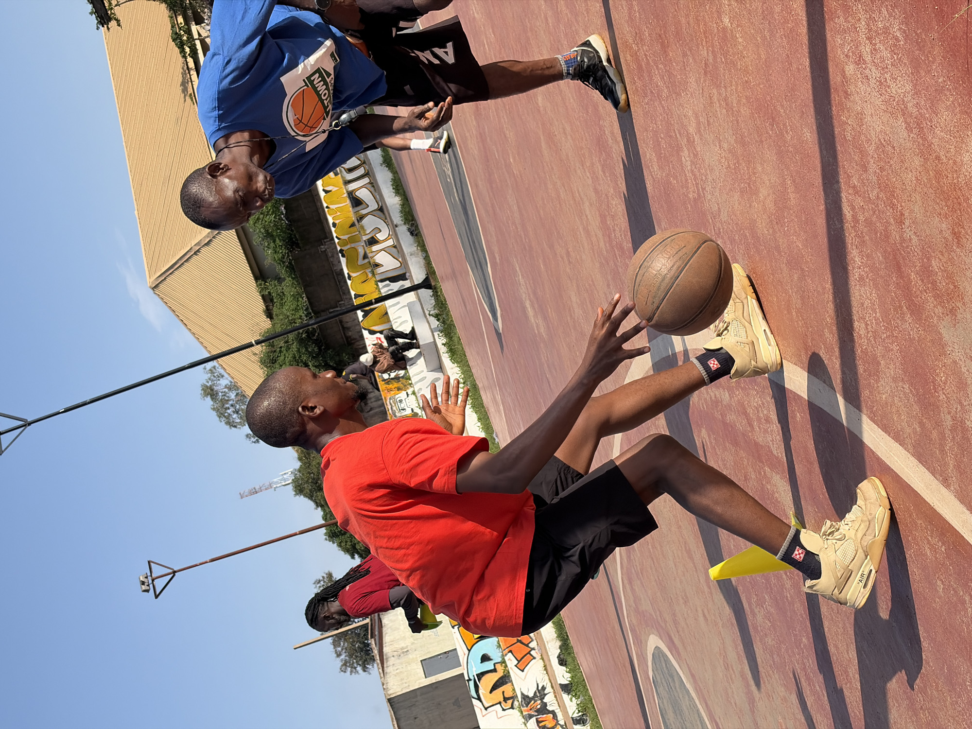 Two adult males are engaged in a basketball drill on an outdoor court. The man in the foreground, wearing a red t-shirt and black shorts, is dribbling a basketball. He has a determined expression and his body is angled forward, indicating focus on the drill. To his right, a man in a blue t-shirt with a "BASKETBALL" graphic and black shorts, appears to be instructing or defending. He is leaning forward, observing the dribbler. The court surface is reddish-brown with white lines. In the background, a building with a light brown, angled roof is visible, along with some graffiti on a wall and a lamppost. The sky is clear and blue. The overall scene suggests an active sports environment. The location is identified as Jos, Nigeria.