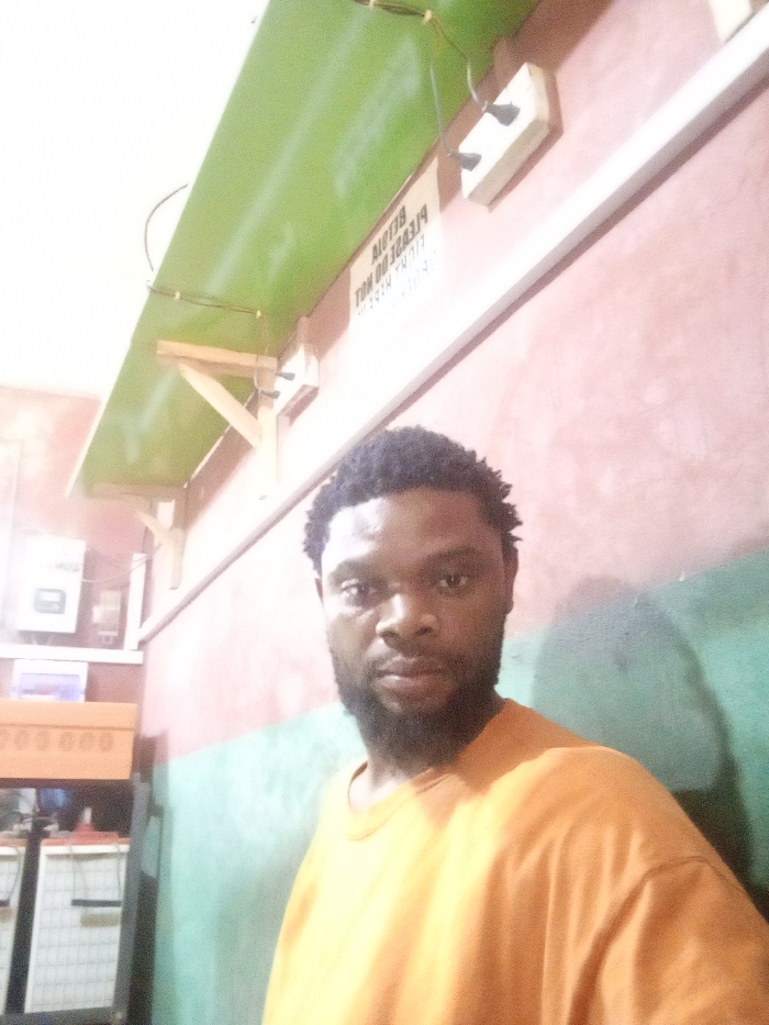 A man with a beard and short, curly hair wears a bright orange t-shirt. He stands inside a room with a color-blocked wall painted pink, then green. A green shelf hangs from the ceiling above him, held up by wooden supports and electrical outlets. A sign is fixed to the wall, illegible due to the angle. A cabinet and batteries are visible on the left side of the frame.
