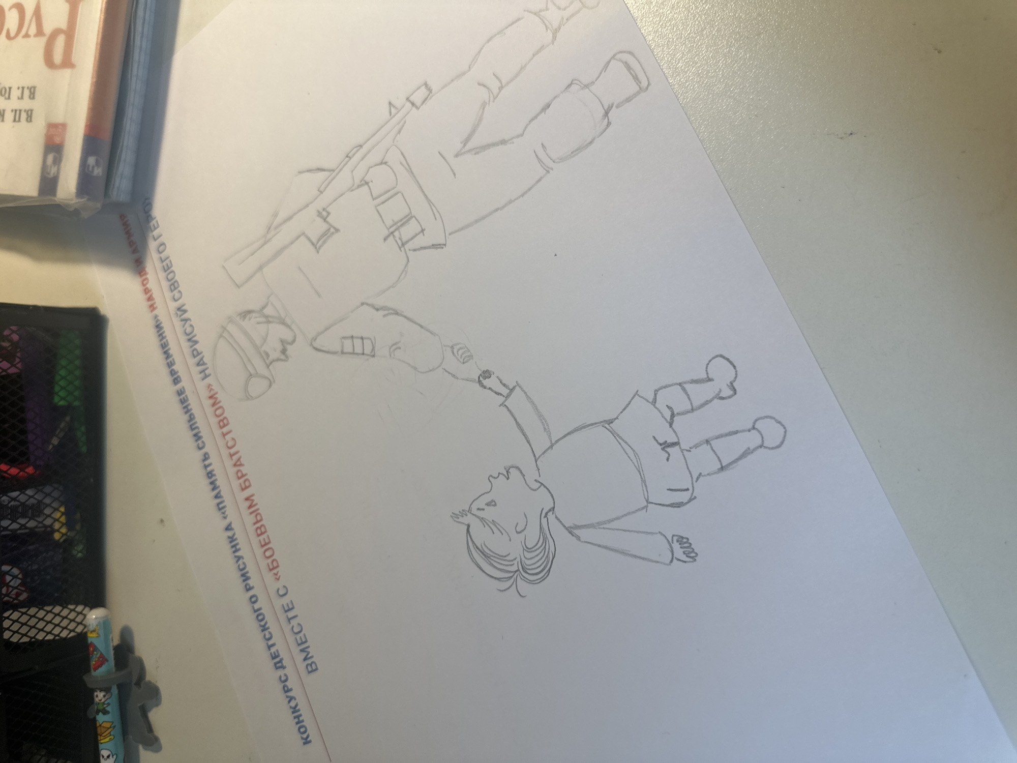 The image shows a pencil drawing on a white piece of paper. The drawing depicts two figures: one appears to be a soldier with a rifle and uniform, and the other appears to be a child. The figures are sketched in a simple, cartoonish style. The drawing is titled "Together with 'Combat Brotherhood' draw your hero" The paper is placed on a surface next to a book and a pen organizer. The printed text along the edge of the paper suggests the drawing is part of a children's drawing contest called "Memory is Stronger Than Time". The location in Moscow, Russia is not identifiable from the image.
