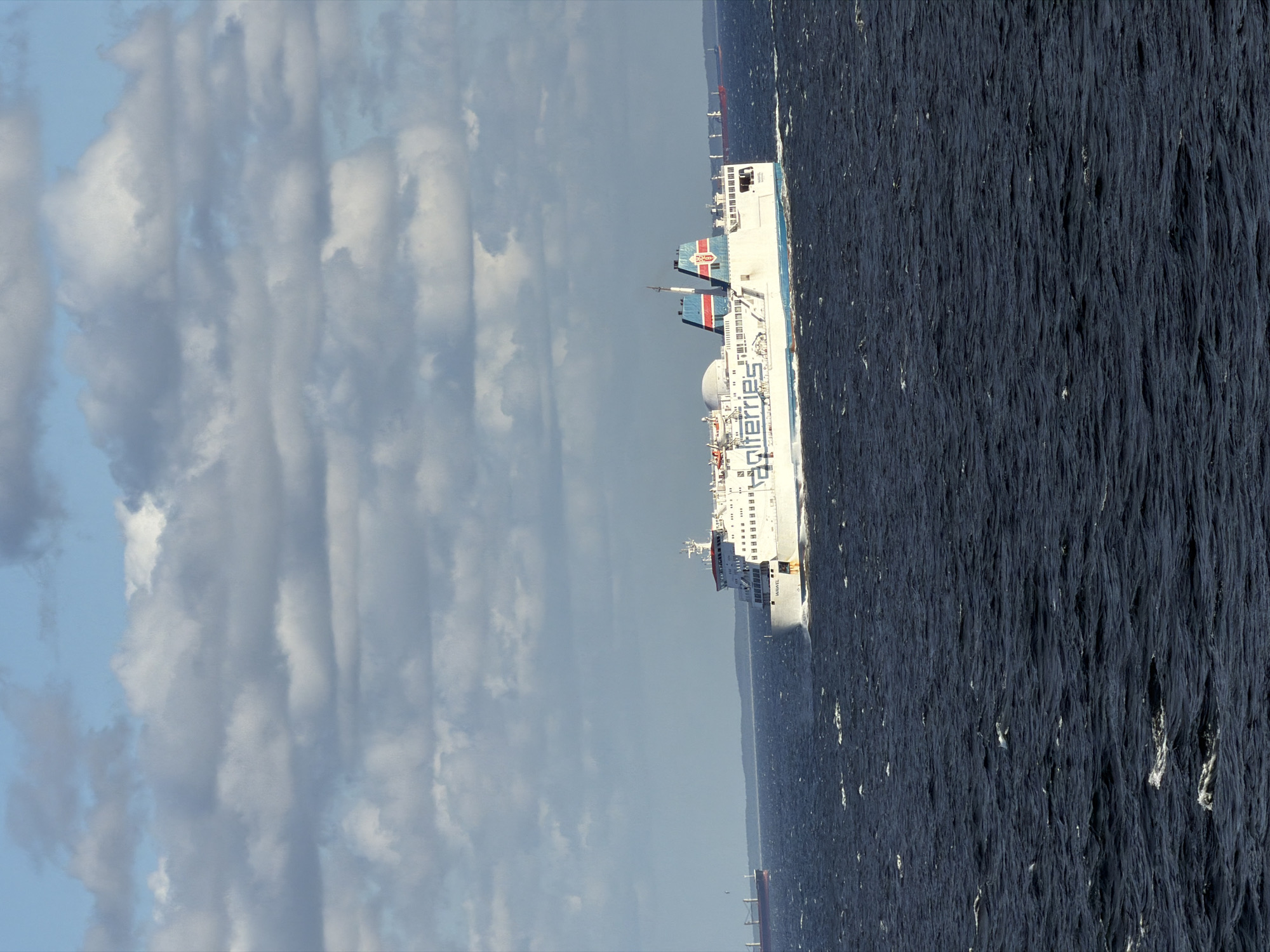An expansive, choppy, dark blue sea fills the bottom two-thirds of the frame, reflecting a cloudy, light blue sky. A large, white ferry with blue trim cuts through the water on the right side of the image, leaving a white wake. The ferry has the word "Stena" in blue lettering on its side, followed by a logo with red and blue stripes. In the distance, on the left, a large, dark red cargo ship is visible against a hazy, green landmass. The sky above is filled with scattered, grey and white clouds. The scene appears to be a wide shot of maritime traffic, possibly in the vicinity of Gdynia, Poland, given the presence of large ferries and cargo ships.