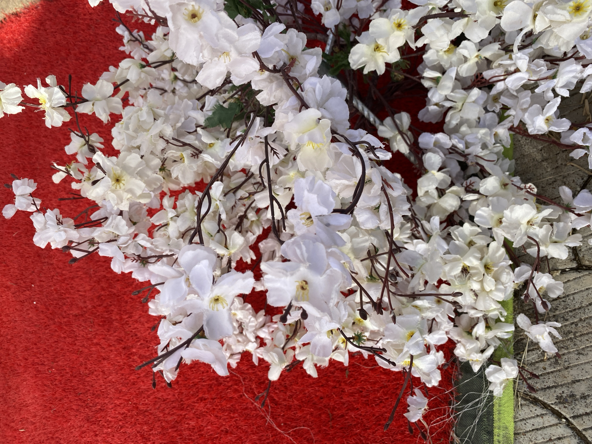 A high-angle shot depicts a dense cluster of artificial white flowers with brown stems, partially covering a red surface on the left and a concrete surface with a strip of green fabric on the right. The scene is brightly lit. No people are visible. The location is specified as Bukuru, Nigeria.