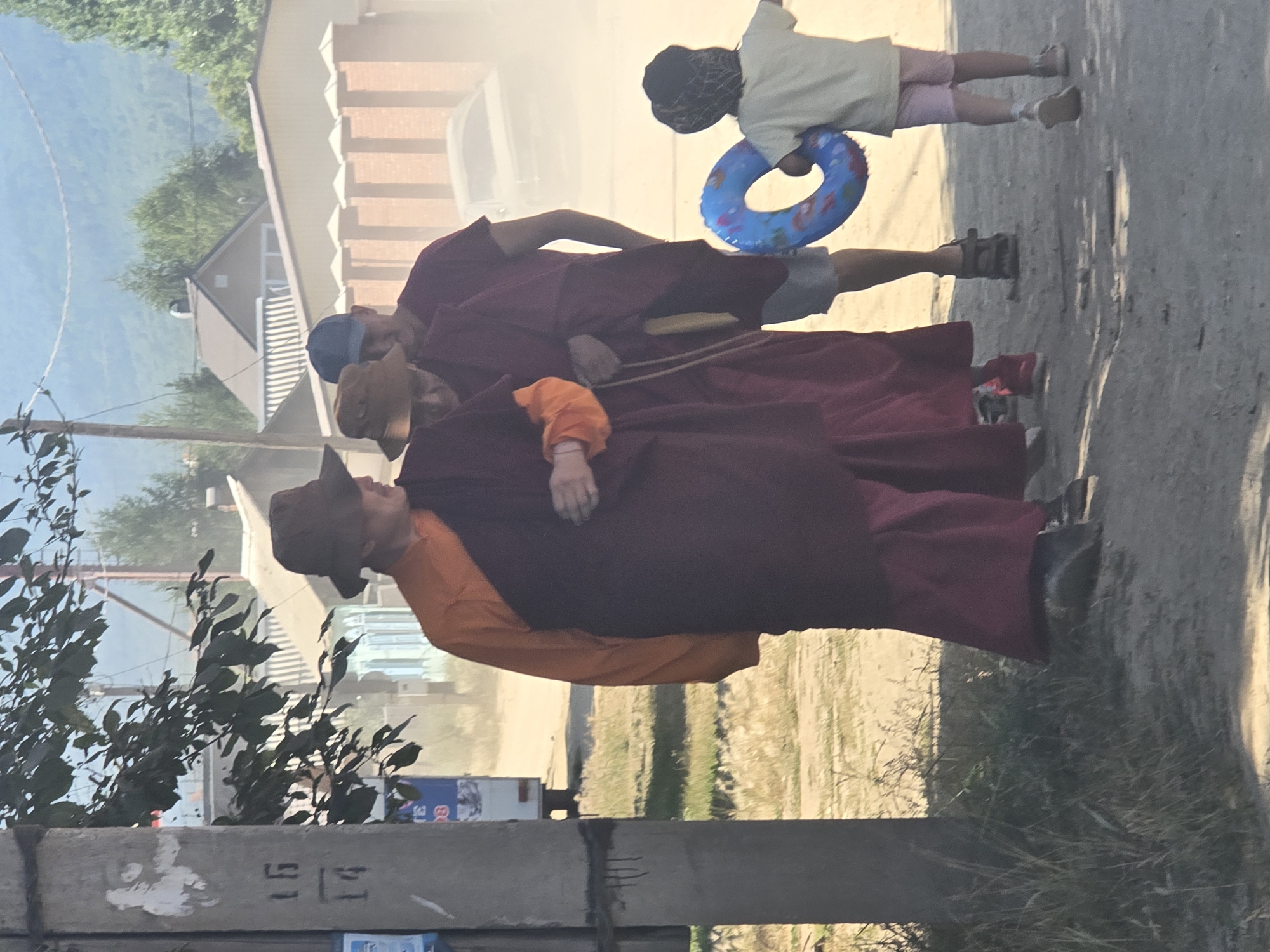 In Maksimiha, Russia, three individuals dressed in maroon robes, two with hats, are walking on a dirt road. The first, on the left, wears an orange undergarment and a dark brown hat. The second, in the middle, wears a lighter brown hat. The third, on the right, wears a blue cap and grey shorts. They are walking away from the viewer, with the first two seeming to be in conversation or interaction. Behind them, a young child in a light yellow t-shirt and pink shorts walks ahead, holding a blue inflatable ring with a fish pattern. In the background, there are houses with fences, a car, and trees, under a hazy sky. A utility pole with markings is visible on the left side of the frame. The ground is covered in dirt and sparse vegetation.