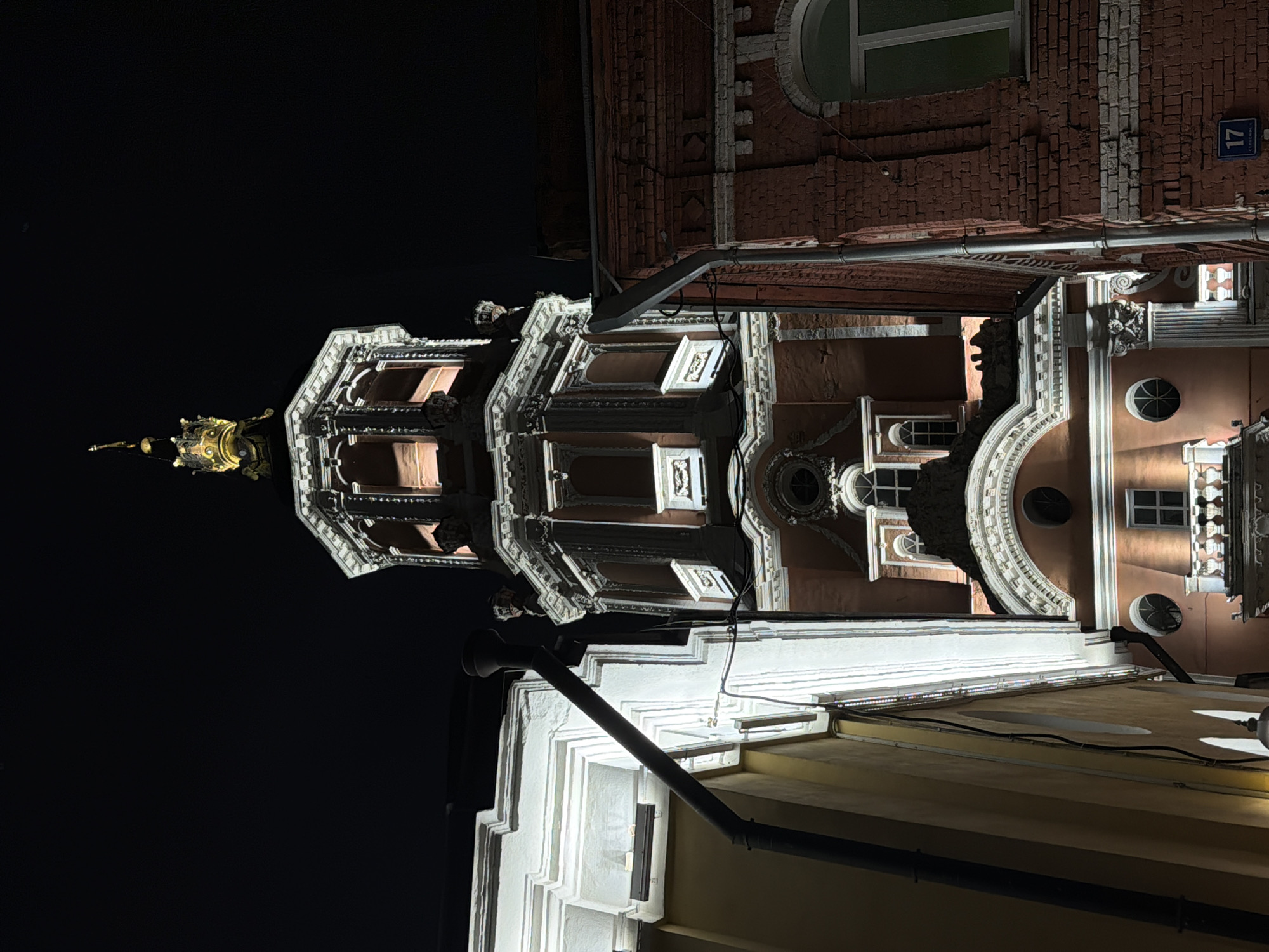 The image, taken at night, shows the Church of the Holy Martyr Antipas on Kolymazhny Dvor, located in Moscow, Russia. The perspective is from below, looking upwards, highlighting the architectural details of the church tower. The top of the tower is adorned with a golden dome. Additional buildings surround the church on the right and bottom of the frame, partially illuminated, while the background is a dark, solid black. A street sign is visible on the right side of the church, displaying "17" in white letters, and Cyrillic text in blue color.