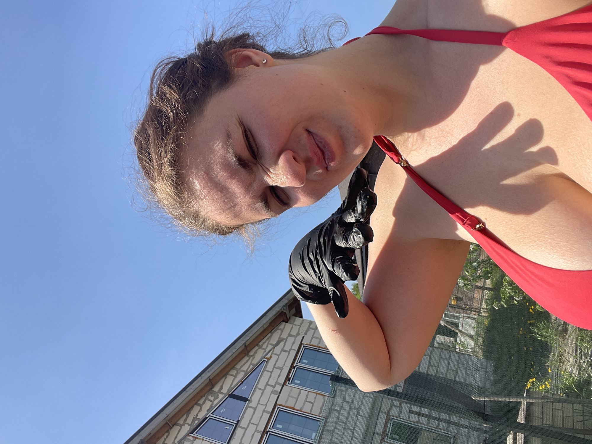 A medium shot from a low angle captures a woman wearing a red bikini and black gloves, with a house and a clear blue sky in the background. The woman's face is tilted downwards and to the right, with her eyes closed and a slight grimace. She is positioned in front of the camera, with the house slightly out of focus in the background, and the sky taking up the upper portion of the image. The house appears to be under construction, built from concrete blocks, with multiple windows visible. The scene is in Мосметаллоконструкция Онт, Russia.