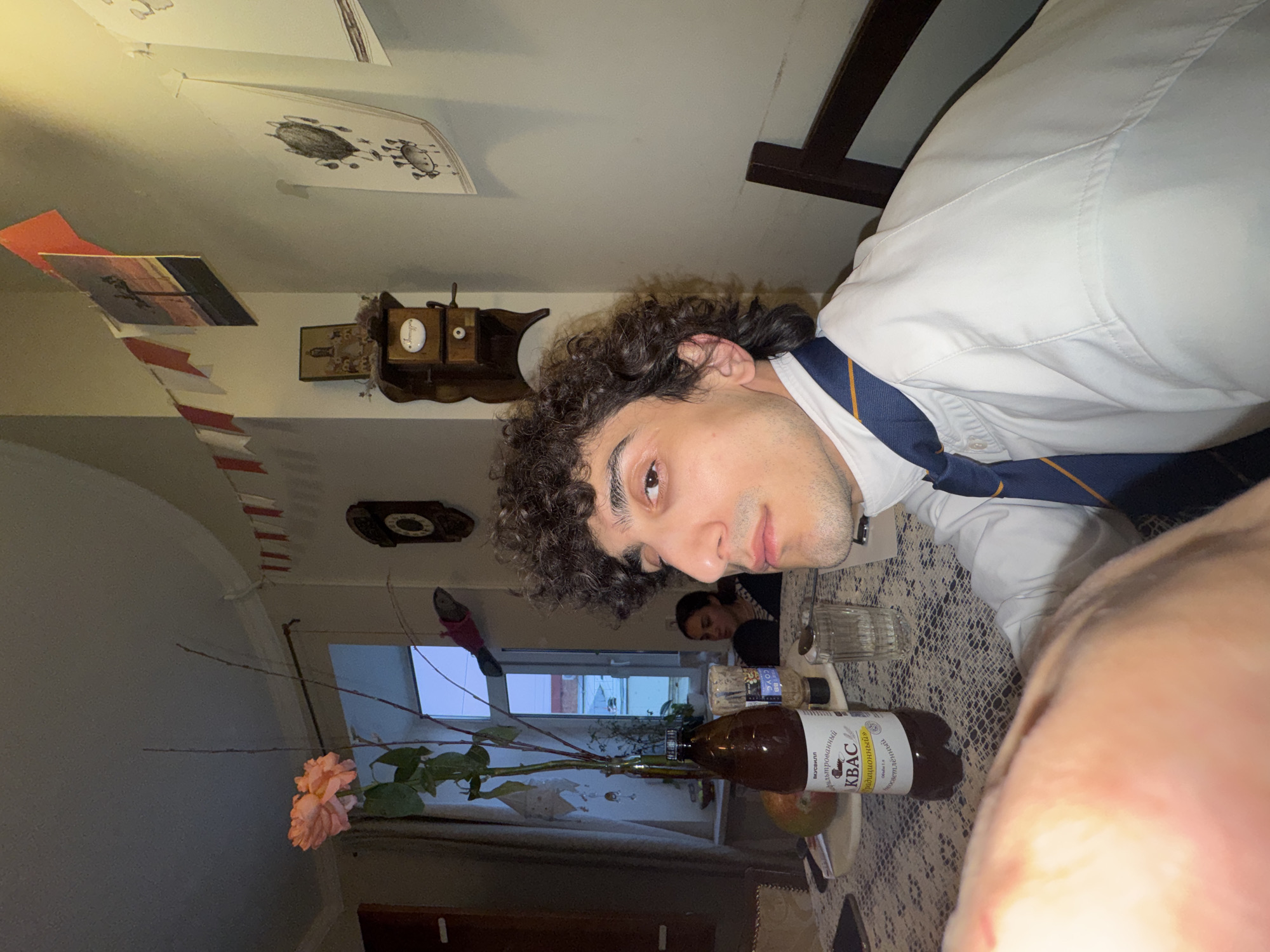 The image shows a man with curly hair wearing a white shirt and a blue and gold striped tie, seemingly sitting at a table in a room. There is a bottle of "Kvass Traditional" on the table, along with other containers, a glass, and what appears to be a rose in a vase. Through the window, a view of what appears to be an urban landscape is visible. Given the "Kvass" bottle, it is plausible the location is in Russia, potentially Moscow.
Another person is barely visible in the background. Various decorative items adorn the wall, and papers are hung from the ceiling. The table is covered with a lace-patterned tablecloth.