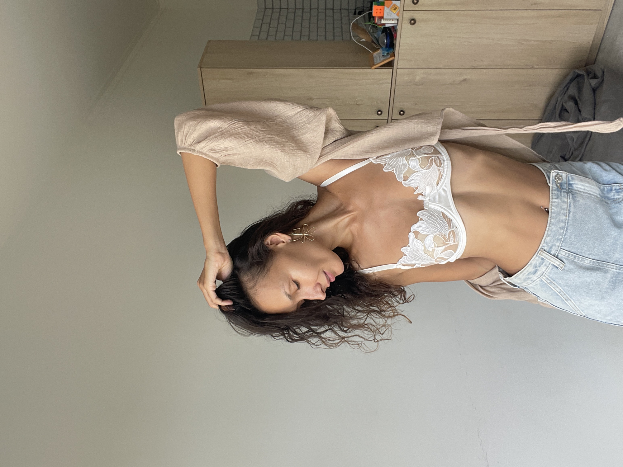 The image depicts a young woman with curly dark hair, wearing a white lace bralette and light blue denim shorts. She is posing with her hand in her hair, looking down and to the side. A light-colored, flowing garment is draped over her shoulders. Behind her, there are light-colored cabinets and a grey beanbag chair. The background is a plain wall. No specific landmarks of Phu Thuy Ward, Vietnam are visible.