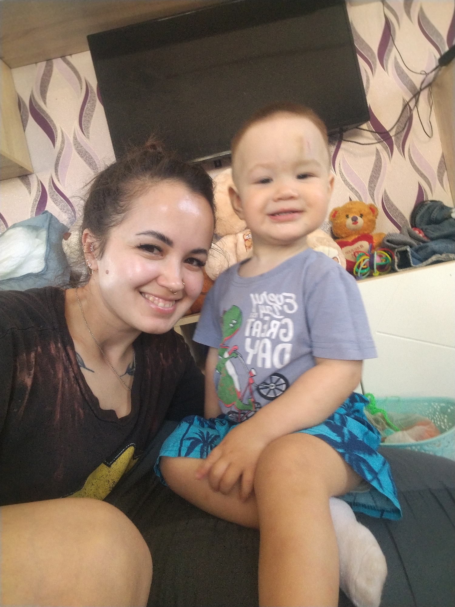 A fair-skinned woman with dark hair pulled back in a bun is smiling at the camera, seated closely beside a fair-skinned young boy with a visible bruise on his forehead. The woman is wearing a dark-colored t-shirt with a graphic on it, and a delicate necklace. The boy is wearing a light blue t-shirt with a dinosaur design and blue shorts with a palm tree pattern. He is also smiling. They are seated on a dark-colored surface.
Behind them is a white shelf on which stuffed animals and other items are placed. A flat-screen television is mounted on the wall. The wallpaper features a pattern of vertical stripes in light and dark shades, possibly in the location of Unknown, Unknown.