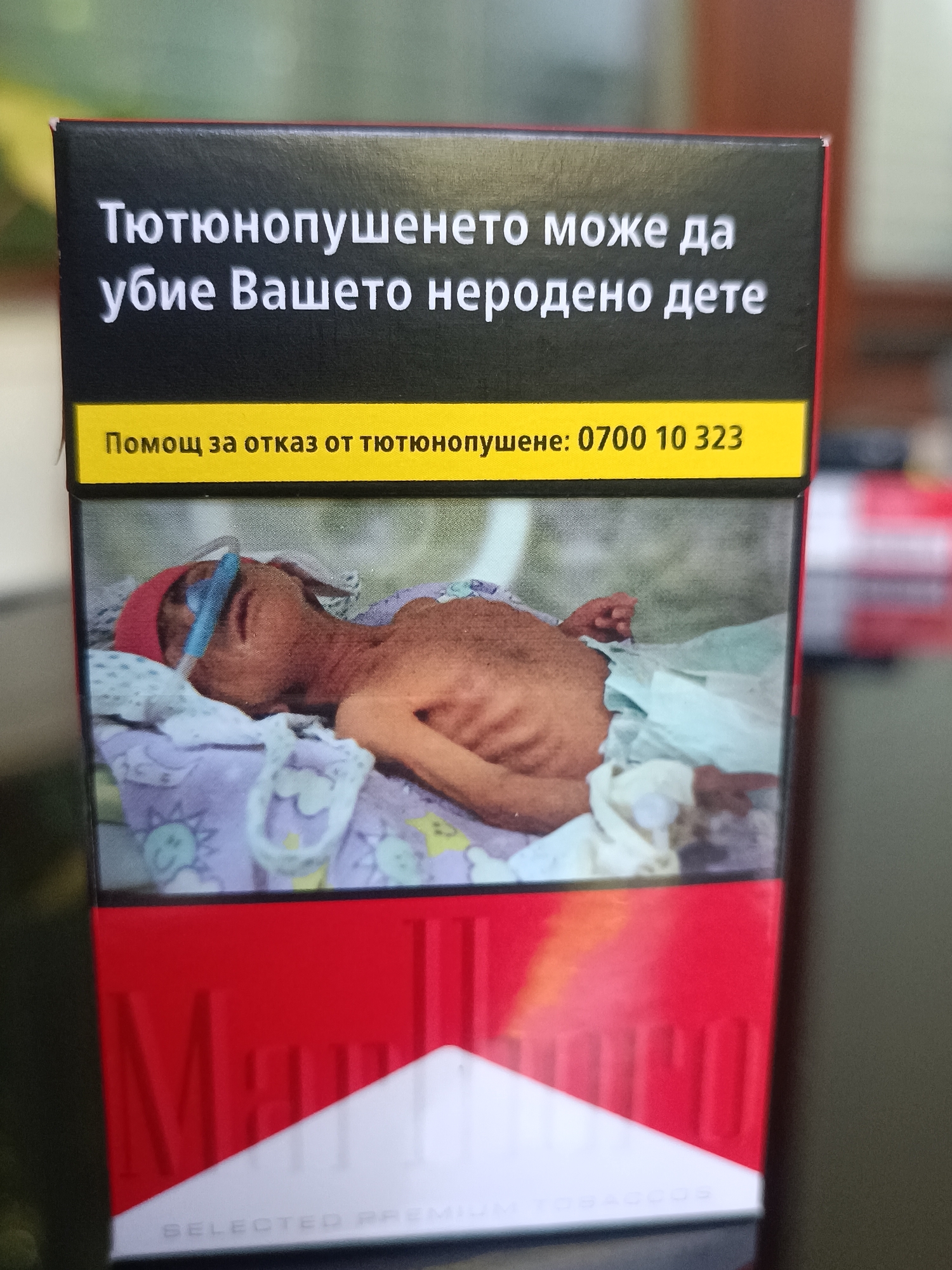 A pack of Marlboro cigarettes is pictured. The pack is red and white, with the Marlboro logo prominently displayed. The upper portion of the pack features a stark warning in Bulgarian: "Smoking can kill your unborn child." Below the warning, a yellow band displays a helpline number for smoking cessation. The center of the pack shows a disturbing image of a very thin, emaciated infant lying in a crib, connected to medical equipment, with a nasogastric tube inserted. The infant appears to be suffering from severe malnutrition or illness. The background of the image is blurred and appears to be an outdoor environment, possibly a garden or park. The overall presentation is intended to convey the severe health risks associated with smoking, particularly to fetuses and infants.