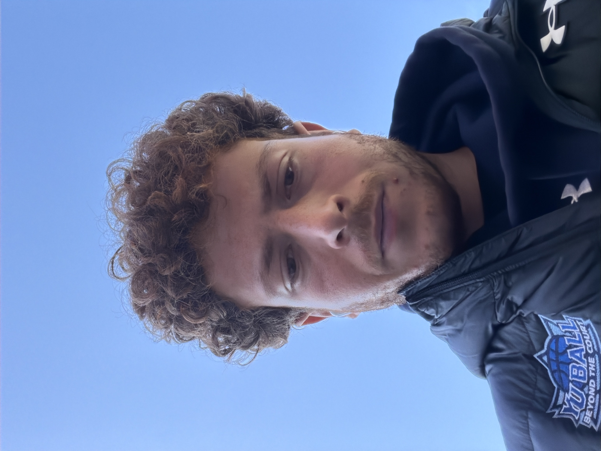 A person is looking directly down at the camera. The person has curly brown hair, light skin, and a short beard. They are wearing a black hooded jacket with a white logo on the right breast. The background is a clear blue sky. The image does not provide enough information to determine the exact location within New York City or specific actions or interactions taking place.