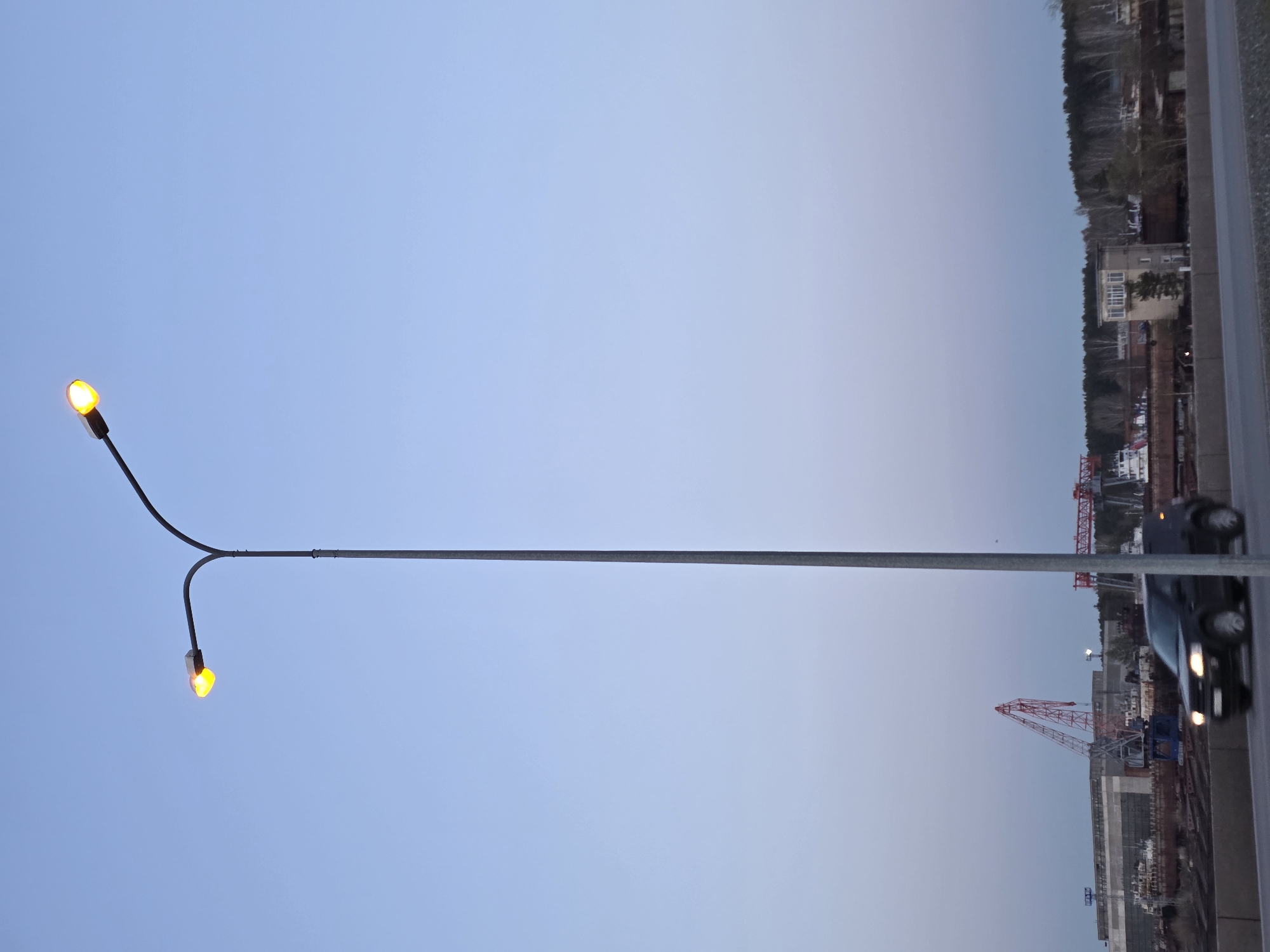 The image shows a streetlamp with two glowing bulbs against a light blue and white gradient sky. In the background, there is a view of a city including cranes, industrial areas, buildings, and a treeline. There is also a car passing by on a road, slightly blurred. The location is likely Chaikovsky, Russia, as suggested in the prompt.