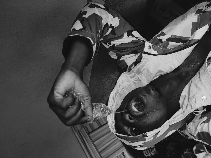 A black and white photograph, captured from a low-angle perspective, depicts a dark-skinned male subject in an indoor setting within Udu, Nigeria. The individual wears a hooded camouflage-patterned jacket, partially unzipped, with the hood covering his head. He is actively pouring liquid from a clear plastic sachet, held in his right hand above his head, directly into his wide-open mouth. His head is tilted sharply backward, and streams of water are visible entering his mouth, spilling down his chin, and wetting his neck. The immediate background shows a plain, textured wall with minor imperfections. A dark surface or fabric is positioned behind his upper back. On the far left, a vertically striped pattern is partially visible, and in the lower right background, the word "GIVENCHY" is faintly discernible on a dark object.