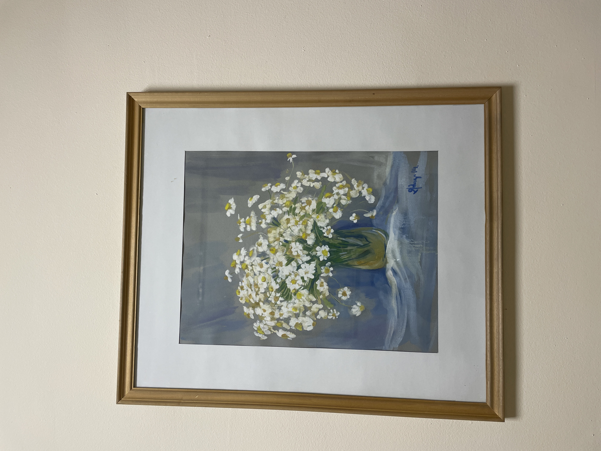 A framed still life painting is centrally displayed on a textured, light beige wall within an indoor setting in Yerevan, Armenia. The artwork, appearing to be a watercolor or thin acrylic, depicts a bountiful bouquet of white and yellow daisies with green stems. These flowers are arranged in a translucent, greenish-yellow glass vase, which sits on a light blue draped surface against a muted blue-grey background. A blue signature is present in the lower right corner of the painting. The artwork is contained within a white mat and a light brown wooden frame with a simple, rounded profile. No individuals or specific actions are depicted in the scene.