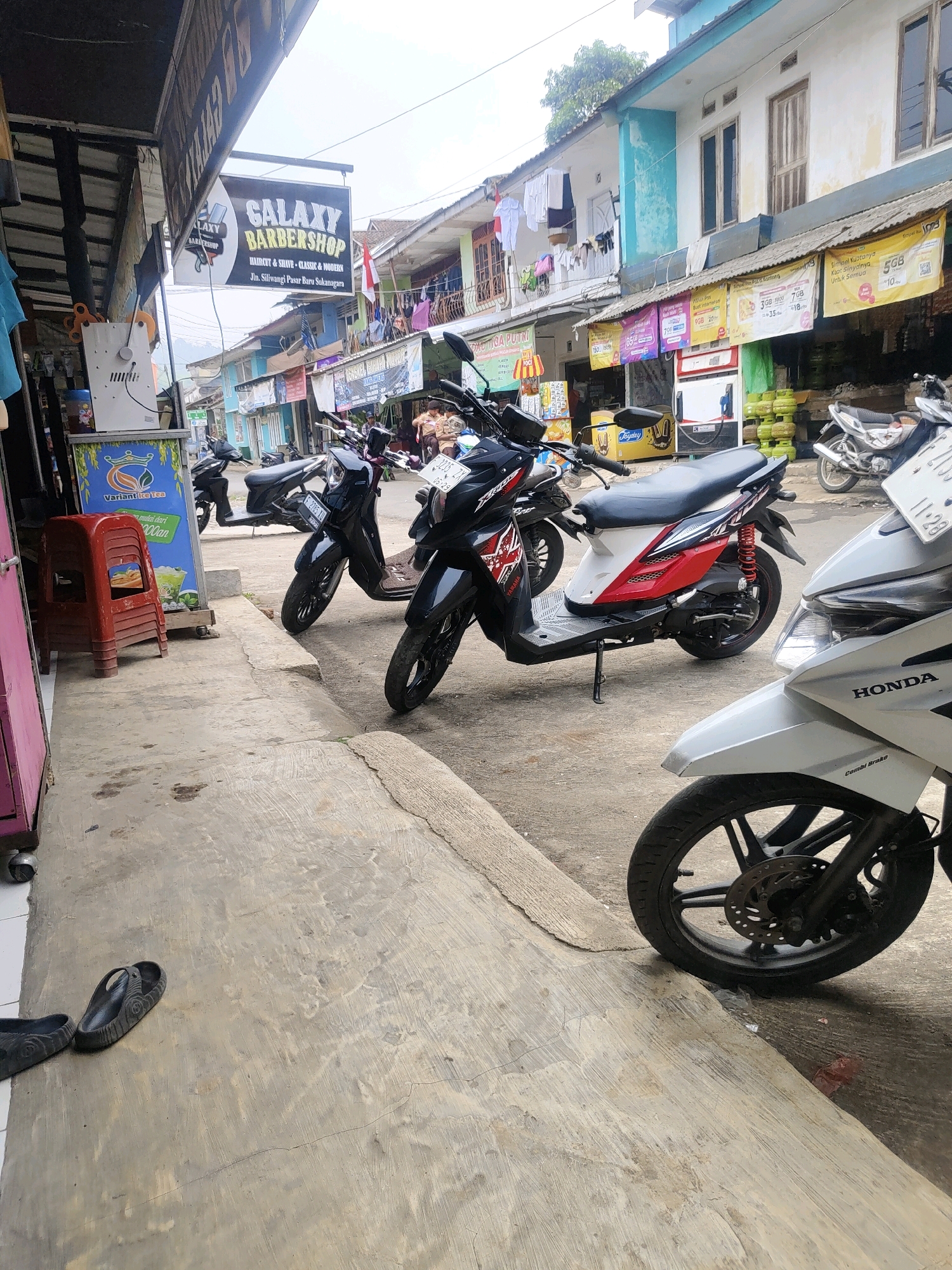 This image depicts a street scene in Sukanagara, Indonesia, with a focus on a barbershop and parked motorcycles. A prominent sign for "GALAXY BARBERSHOP" is visible, indicating services like "HAIRCUT & SHAVE - CLASSIC & MODERN" and an address on "Jl. Siliwangi Pasar Baru Sukanagara". Several motorcycles are parked along the roadside, including a white Honda in the foreground and black scooters further down the street. The environment appears to be a commercial area with shops and residential buildings lining the street. Laundry is hung out to dry on a balcony in the background. The ground is paved with concrete, and there are some scattered items on the sidewalk, including a red plastic stool and a pair of black flip-flops. The overall scene suggests a typical day in a bustling Indonesian town.
