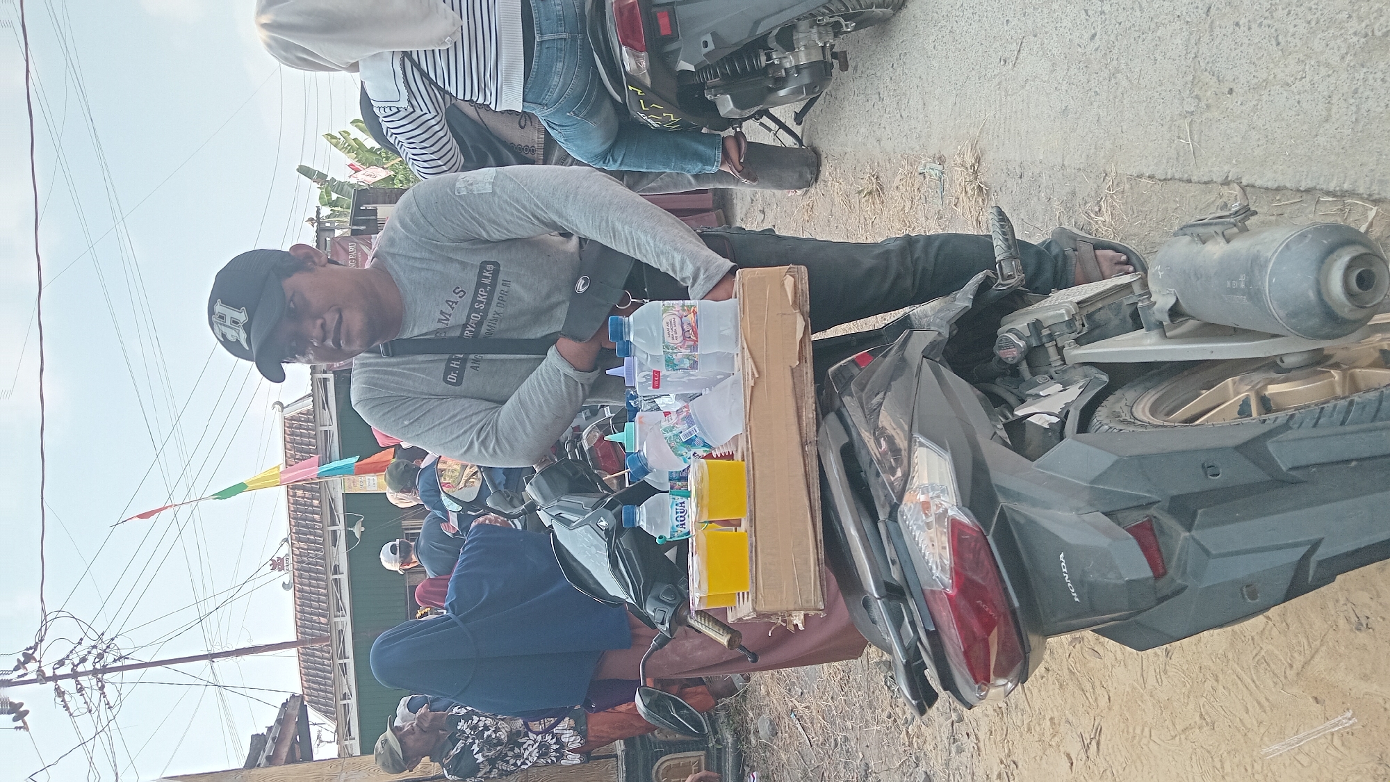 In Kedungwaru, Indonesia, a man wearing a grey long-sleeved shirt with writing on it and a black baseball cap stands next to a motorcycle. He is holding a cardboard tray containing four plastic cups filled with yellow liquid and several bottles of water. The motorcycle is parked on a dirt and concrete surface. In the background, several other people are visible, some wearing headscarves, and a colorful banner is hanging from a pole. There are also power lines and houses in the background.