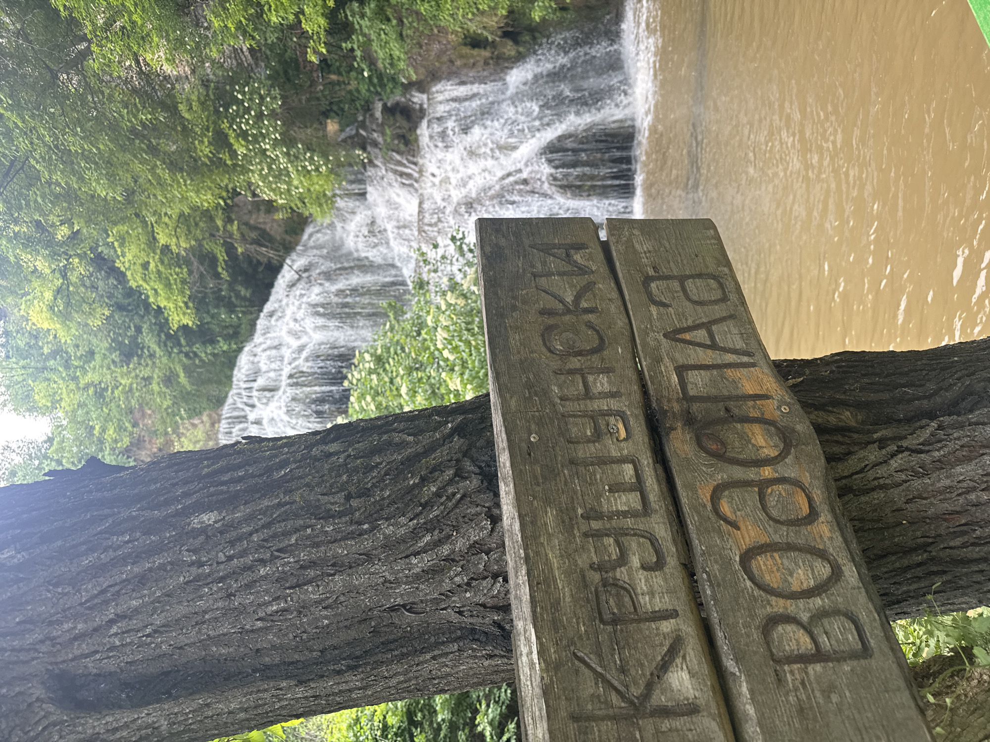 A close-up shot of a wooden sign in Krushuna, Bulgaria, with the Cyrillic inscription "Крушунски Водопад" (Krushuna Waterfall). The sign rests on a rough-barked tree branch. A cascading waterfall is visible in the background, with water flowing into a brown river. Green trees are seen on the left side of the frame.