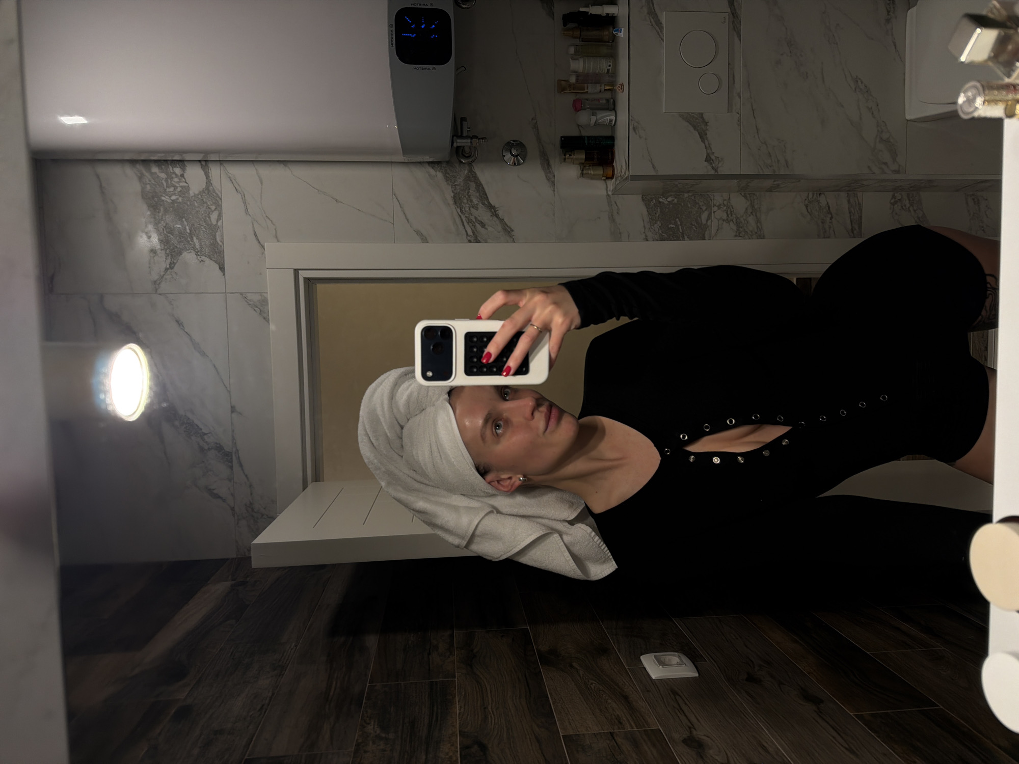 A female individual is depicted taking a mirror selfie in a private bathroom, located within a residence in Budva, Montenegro. The subject, a young adult with light skin, appears without makeup and has her hair wrapped in a white towel, turban-style. She wears a long-sleeved, black, form-fitting bodysuit or romper with a V-neckline secured by a row of unfastened silver-colored snap buttons, exposing a portion of her chest. A tattoo is visible on her upper left thigh. Her right hand, with red-painted fingernails and a ring on the fourth finger, holds a white smartphone with a distinct black and white case, pointed towards the mirror.
The bathroom features two distinct wall tiling patterns: dark wood-grain-effect tiles on the left and white marble-effect tiles with grey veining on the right. A single, bright overhead light source is reflected in the mirror. On the right-hand wall, a white Ariston electric water heater with a blue digital display is mounted horizontally. Below it, a white shelf or vanity top holds an assortment of small bottles, tubes, and cosmetic containers. A white wall-mounted toilet flush plate with two circular buttons is partially visible beneath the shelf. A white electrical outlet is present on the dark wood-grain tiled wall to the left. The bottom edge of the mirror reflects a white countertop in the foreground with a few indistinct objects. A white door frame and a light-colored door are visible in the reflection behind the subject.