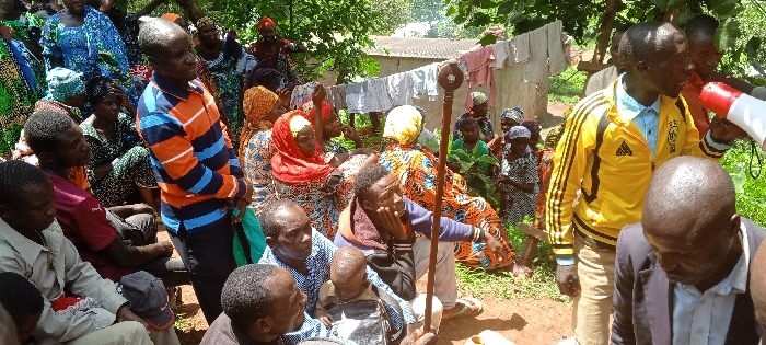 A gathering of people, primarily African, in an outdoor setting. Some are seated, others stand. Many are wearing vibrant, patterned clothing. One man holds a staff, while another uses a megaphone. Clothes hang on a line in the background. The atmosphere seems attentive, perhaps a community meeting or an announcement.