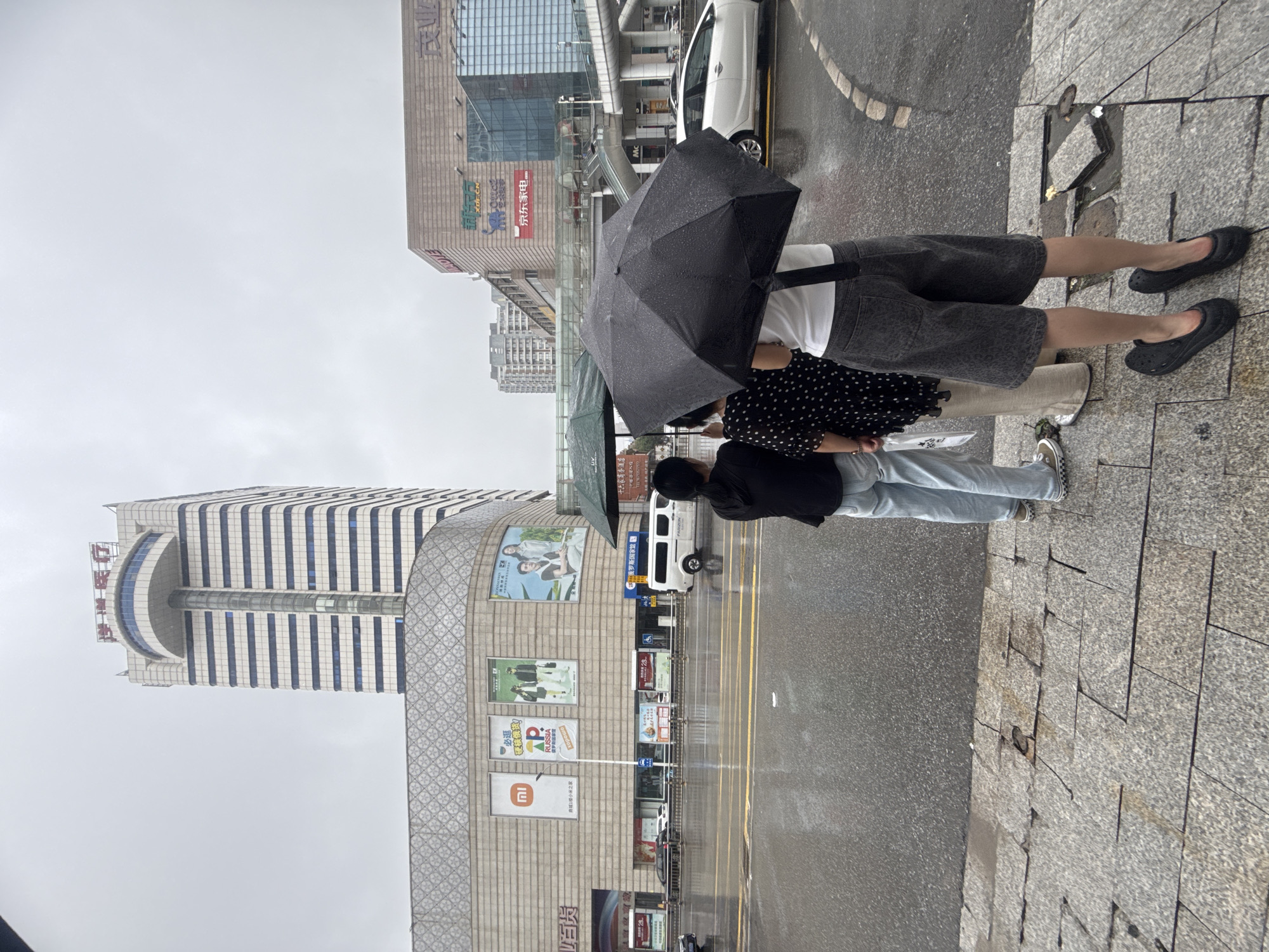 The image shows a street scene in Qinhuangdao, China, on a rainy day. Three people are standing on a sidewalk, waiting under an umbrella. A tall, modern building with signs in Chinese is visible in the background, along with other commercial buildings and vehicles on the wet street. The sidewalk is made of stone tiles, some of which are damaged. The sky is overcast, indicating rainy weather.
