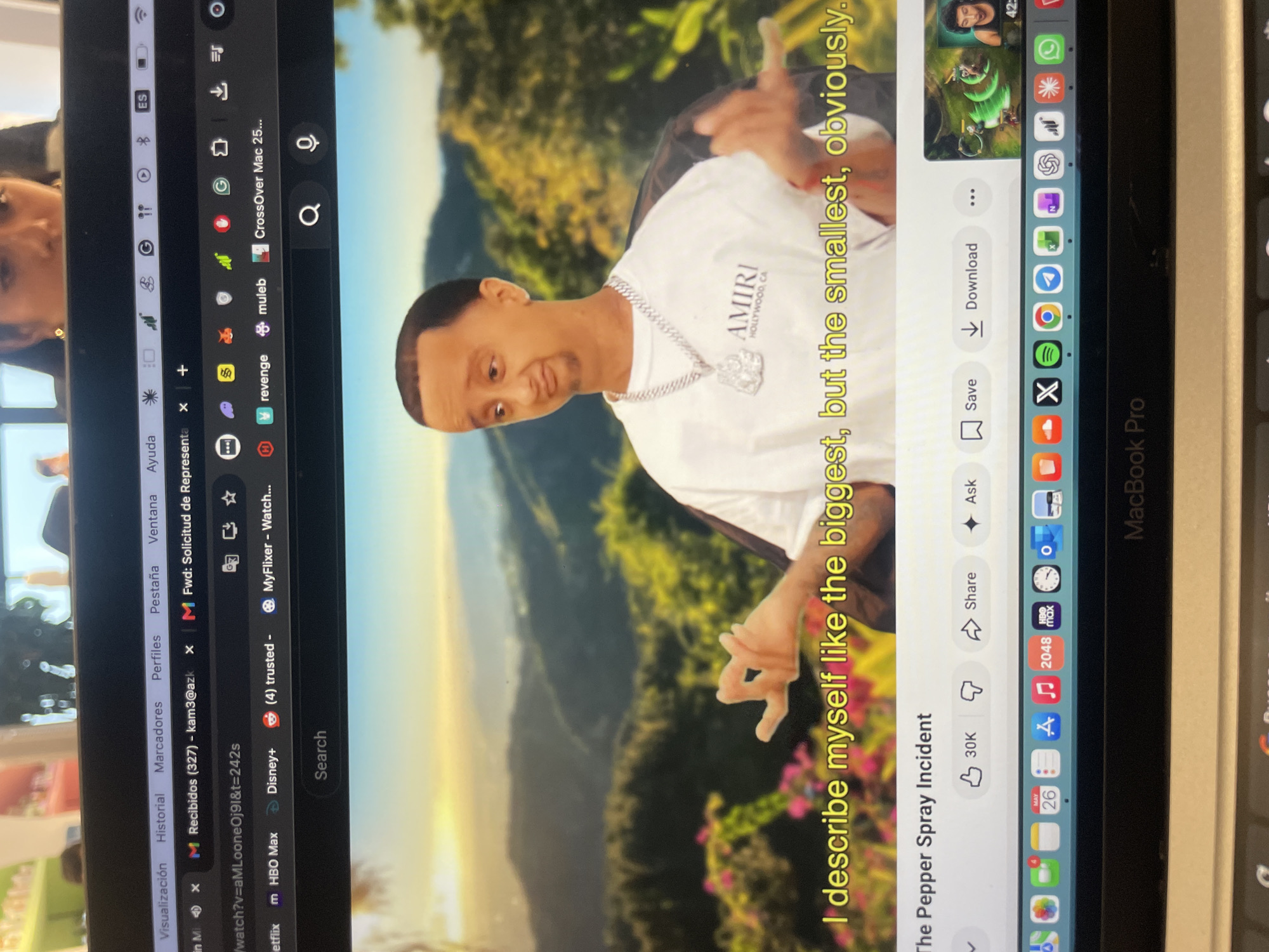 A photograph shows a MacBook Pro screen displaying a YouTube video of a man wearing an "AMIRI" shirt and a silver chain, stating, "I describe myself like the biggest, but the smallest, obviously," titled "The Pepper Spray Incident." The MacBook Pro's dock is visible with various app icons, and several open browser tabs are also shown, including email, streaming services (Netflix, HBO Max, Disney+), and a file related to a representation request. The operating system is set to Spanish.