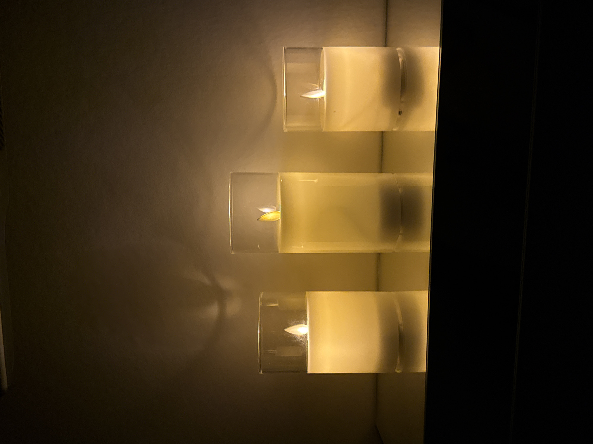 Three led candles in glass cylinders are arranged on a reflective surface. The candles emit a warm, yellow light and cast shadows on the wall behind them. The overall lighting is dim, creating a cozy and atmospheric scene. The image is taken indoors, and the background is a neutral, textured wall. The location in Votkinsk, Russia is not visible in the image.