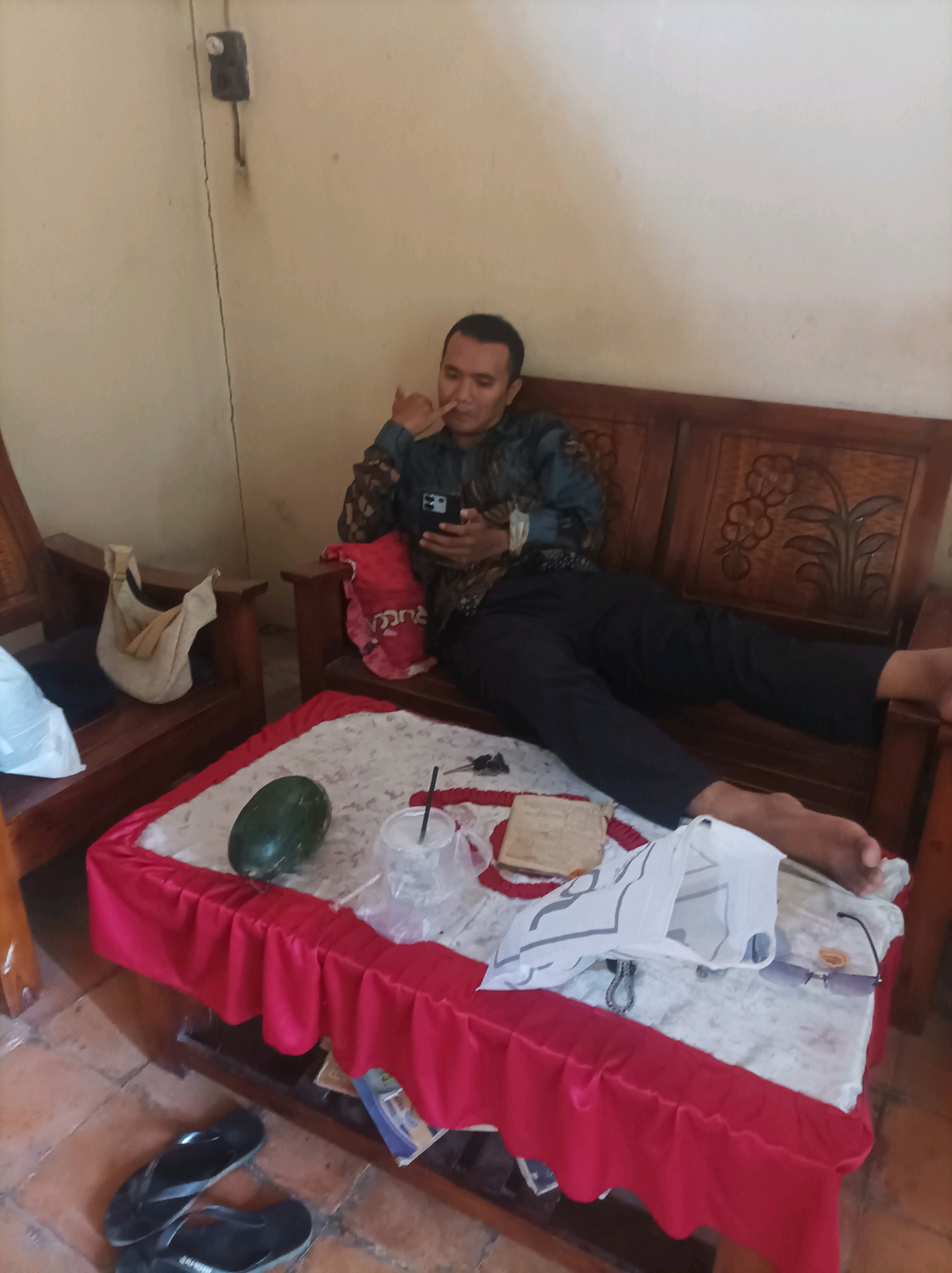 The image depicts an indoor scene, likely a living room, with a man seated on a wooden sofa. The man is wearing a dark patterned shirt and dark trousers, and he is looking at his phone, with his right hand held to his face. A coffee table in front of the sofa is covered with a red tablecloth that has a ruffled edge. On the table, there is a watermelon, a clear plastic cup with a straw, a small stack of papers or books, and a white bag with a grey pattern. A pair of black flip-flops are on the floor to the left of the table. The walls are a light color, and there is a power outlet visible on the left wall. The overall impression is a casual, domestic setting. The image is located in Canga'an, Indonesia.