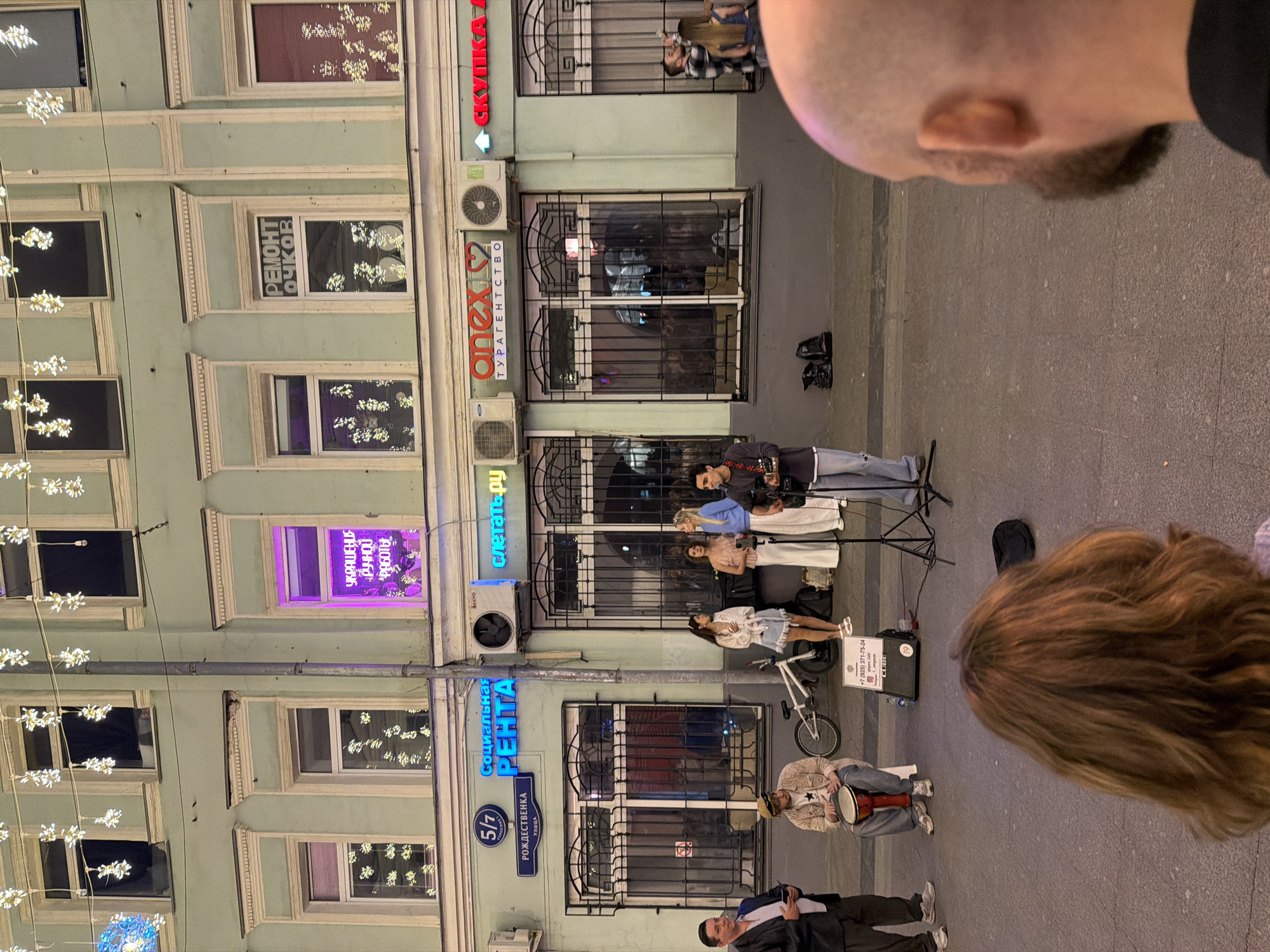 Here is a description of the image:
**Scene:** A vertical outdoor shot, slightly askew. The scene depicts a Moscow street in Russia. A musical street performance is taking place.
**Environment:** The architectural backdrop consists of a multi-story building with a pale green facade and decorative window frames. There are shops or business establishments on the ground floor, indicated by illuminated signs and window displays. Decorative lights dangle from above, providing a festive atmosphere.
**Subjects:** Several people are present, likely street performers. There is a band comprised of three women and one man singing in harmony with guitars. They are performing for a crowd.
**Objects:** A bicycle stands behind one of the performers, along with musical instruments, a speaker, and a donation box. Street signs and business signage are visible on the building.
**Actions:** Performers are playing music and singing, while pedestrians are watching, and likely interacting with the performance by listening and donating. The band's performance is taking place on the sidewalk of the city street.
**Interactions:** The musicians interact with each other, creating a performance for the audience. Bystanders are engaging with the performance.
**Additional Details:** Signage in Russian reads "РОЖДЕСТВЕНКА УЛИЦА" indicating the location as Rozhdestvenka Street. Other business names such as "Anex", "Слетать.ру" and "Социальная Рента" are present.