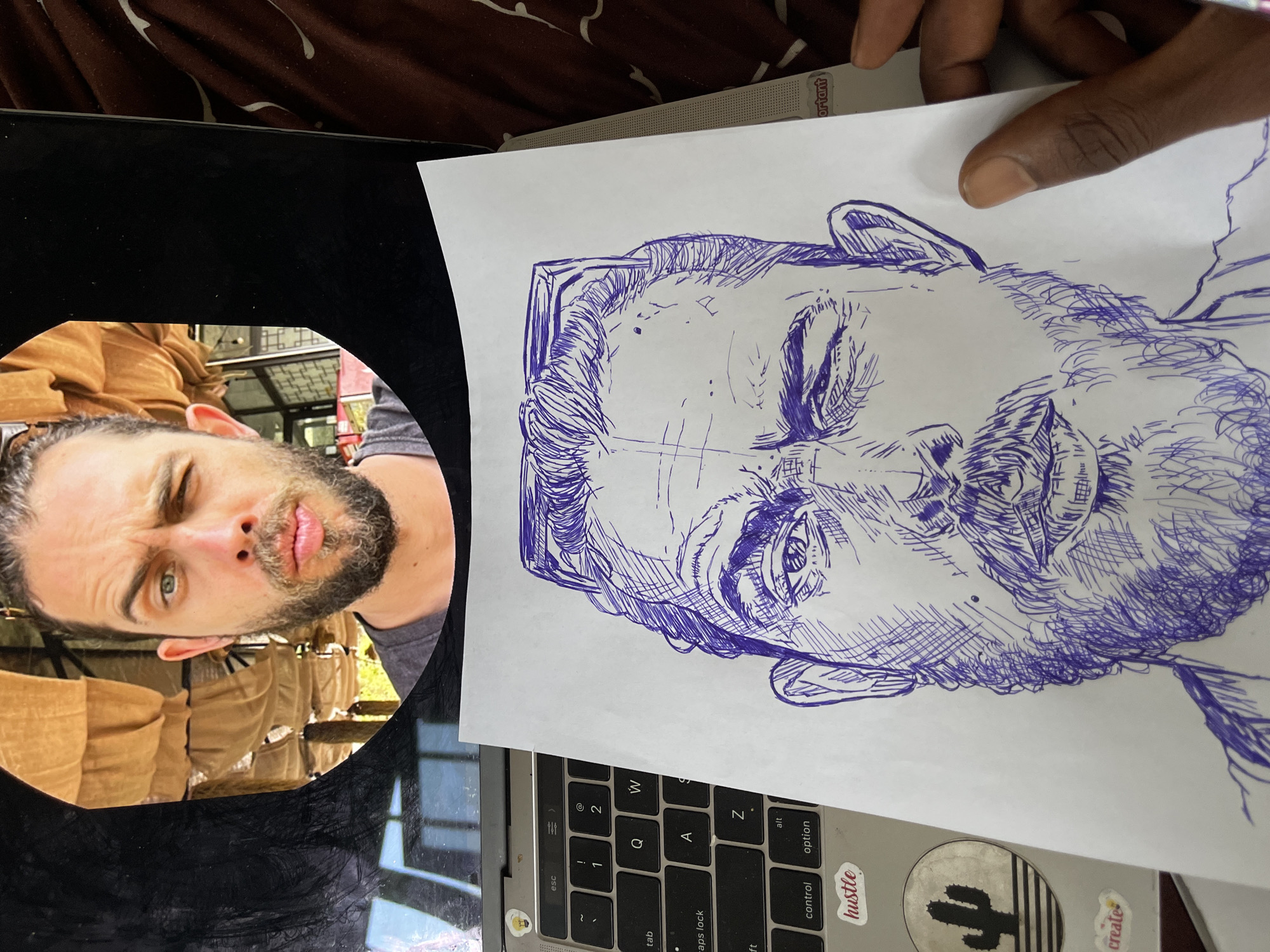 A close-up, overhead shot captures a hand holding a piece of paper featuring a pen sketch of a man's face. The drawing is rendered in blue ink with heavy cross-hatching to create shading and texture, particularly on the beard and hair. The man in the sketch is depicted with one eye closed and the other open, with sunglasses resting on his head.
Behind the paper, the keyboard of a laptop is visible, showing keys like "esc," "tab," "caps lock," "control," and "option." Two stickers are adhered to the laptop: one circular sticker with a cactus design and another small sticker that reads "create." To the right of the laptop, a brown, patterned fabric, possibly a blanket or bedsheet, is visible.
In the upper portion of the image, a circular reflection or screen display shows the face of the same man, appearing to be looking directly at the viewer. He has a beard and his hair is pulled back. His left eye is open and appears to be green, while his right eye is partially closed in a wink. The background of this reflected image shows a blurred scene with fabric drapes and what might be an outdoor setting with trees. The overall impression is of someone working on or reviewing a piece of art, possibly with a reference image. There is no information within the image to indicate the location is Abuja, Nigeria.