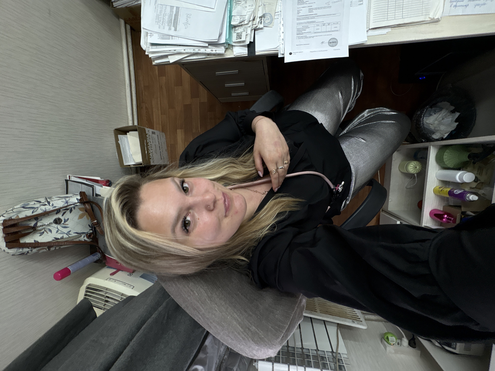 A woman with blonde hair and wearing a black top and silver pants is sitting in a chair in an office. She is looking at the camera. There is a desk with papers on it, and a cabinet with drawers underneath. A trash can is next to the desk. A shelf with various objects is against the wall. A purse and an electric heater are near a wall with vertical lines.
The location is specified as Noviy, Russia, but there are no details in the image that confirm that.