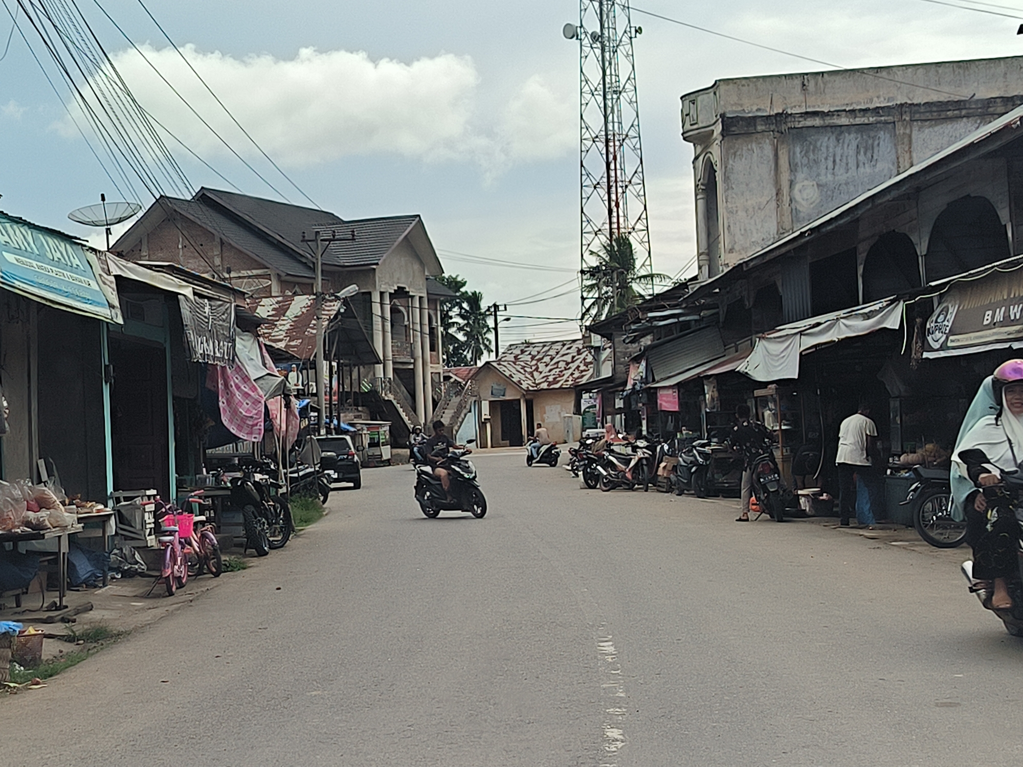 The image depicts a street scene in Cotseutui, Indonesia. The street is paved with asphalt and shows signs of daily use, with some debris on the shoulder. On either side of the street, there are various buildings, some appearing to be shops with awnings and open fronts. The architecture is varied, with some structures having corrugated metal roofs and others appearing more substantial with concrete facades. A prominent feature in the background is a tall, lattice-like telecommunications tower.
Several motorcycles are visible, both parked and in motion, suggesting this is a common mode of transportation. Some people are also present, interacting with the shops or riding the motorcycles. In the foreground on the left, there is a stall or table laden with what appear to be food items or produce. A small pink bicycle is also parked near the stall. The sky is overcast with clouds. The overall impression is a bustling, local street with commercial activity.