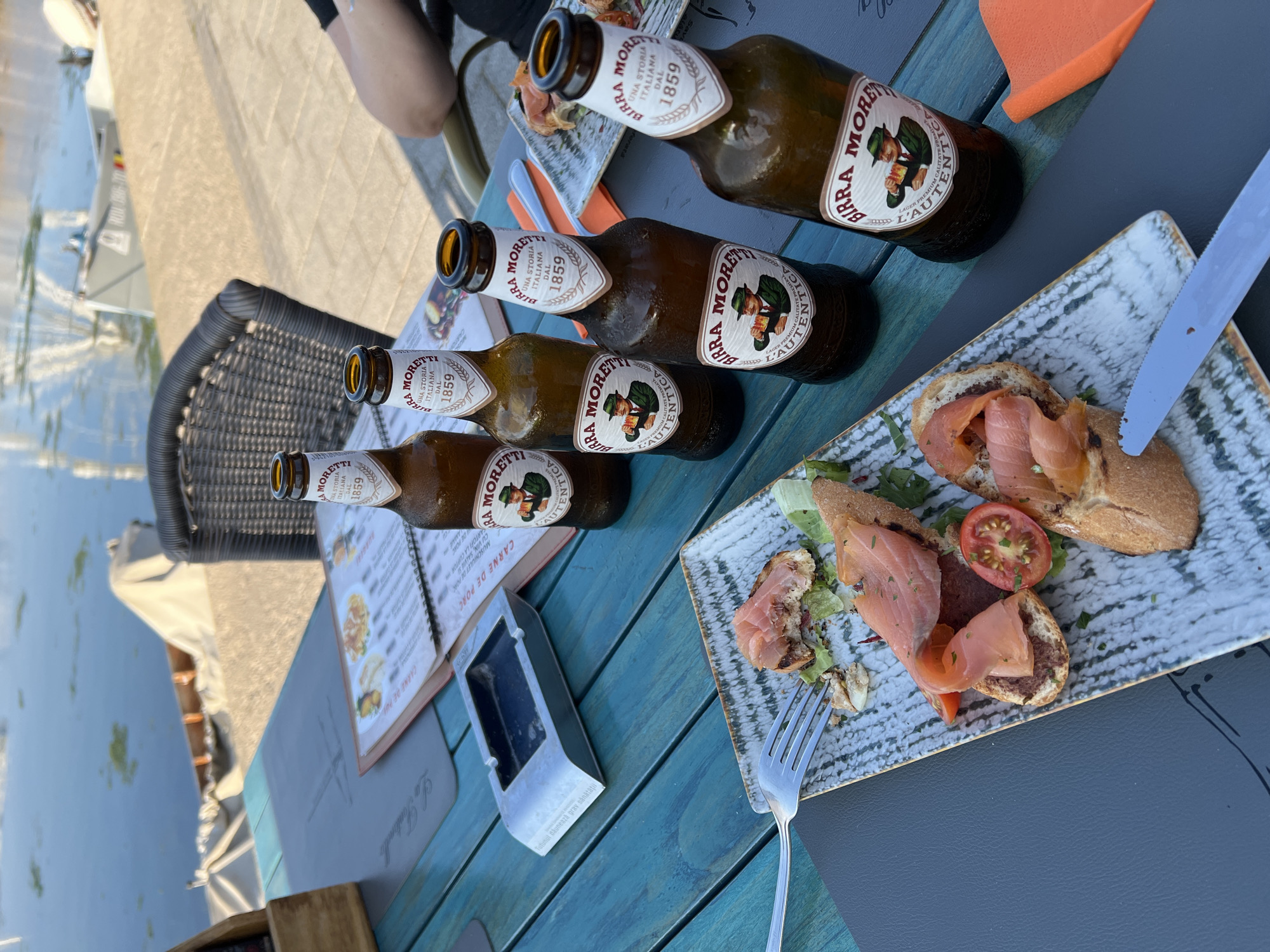 A high-angle medium shot captures a table setting outdoors by a body of water. On the turquoise-colored wooden table are four bottles of Birra Moretti beer, a plate of open-faced sandwiches with smoked salmon and cherry tomatoes, a fork, an ashtray, and an open menu. The background shows a waterfront with boats and a blurred Ferris wheel, suggesting the location is Constanța, Romania, likely along the Tomis Port or Marina. The scene implies a relaxed meal or drink break.