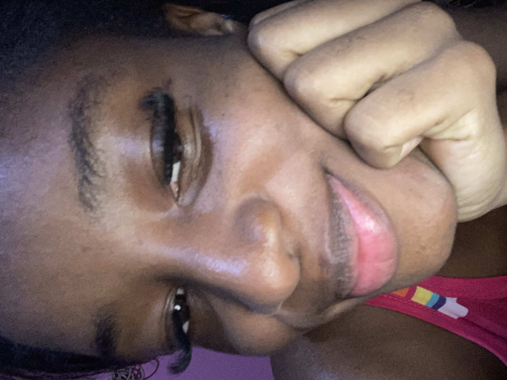 The image is a close-up portrait of a young Black woman. She is resting her chin on her hand, which is curled into a fist. Her eyes are partially closed, and she has a slight smile on her lips. She is wearing a pink top with a colorful striped pattern on the strap. The background is a blurred purple color. There is no discernible information in the image that would indicate a specific location in Jos, Nigeria.