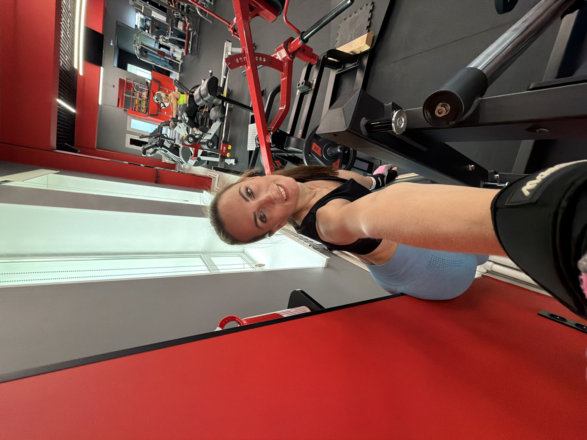 A woman is taking a selfie in a gym in Birobidzhan, Russia. She is wearing a black sports bra and blue leggings. She has her hair pulled back in a ponytail and is wearing small earrings. She is smiling at the camera. In the background, there are various weight machines and exercise equipment. The gym appears to be well-lit and modern, with red and gray accents.