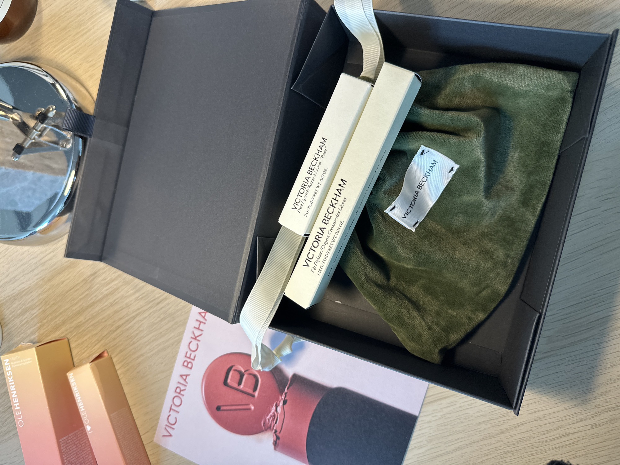 The image displays a collection of cosmetic products and their packaging arranged on a light-colored wooden surface. Central to the scene is an open, black presentation box, revealing its contents. Inside the box, a dark green velvet-like drawstring pouch is visible, featuring a white label embroidered with "VICTORIA BECKHAM". Two small, off-white rectangular product boxes, both labeled "VICTORIA BECKHAM", rest atop the pouch and are connected by a light cream-colored ribbon. One box specifies "POSH LIPSTICK / ROUGE À LÈVRES 'Posh'" with a net weight of "2G / POIDS NET WT. 0.07 OZ.", while the other is marked "LIP DEFINER / CRAYON CONTOUR DES LÈVRES" with a net weight of "1.14 G / POIDS NET WT. 0.04 OZ.". Adjacent to the black box, a 'VICTORIA BECKHAM' branded booklet or magazine features an image of a red lipstick with the initials "VB" embossed on its tip. In the upper left quadrant, two partially visible light orange cardboard boxes from "OLEHENRIKSEN" are present. The background includes a partial view of a reflective metallic object and a dark brown cylindrical item. No individuals are depicted in the scene. The precise location within the city of London, United Kingdom, is not discernible from the visual content of the image.