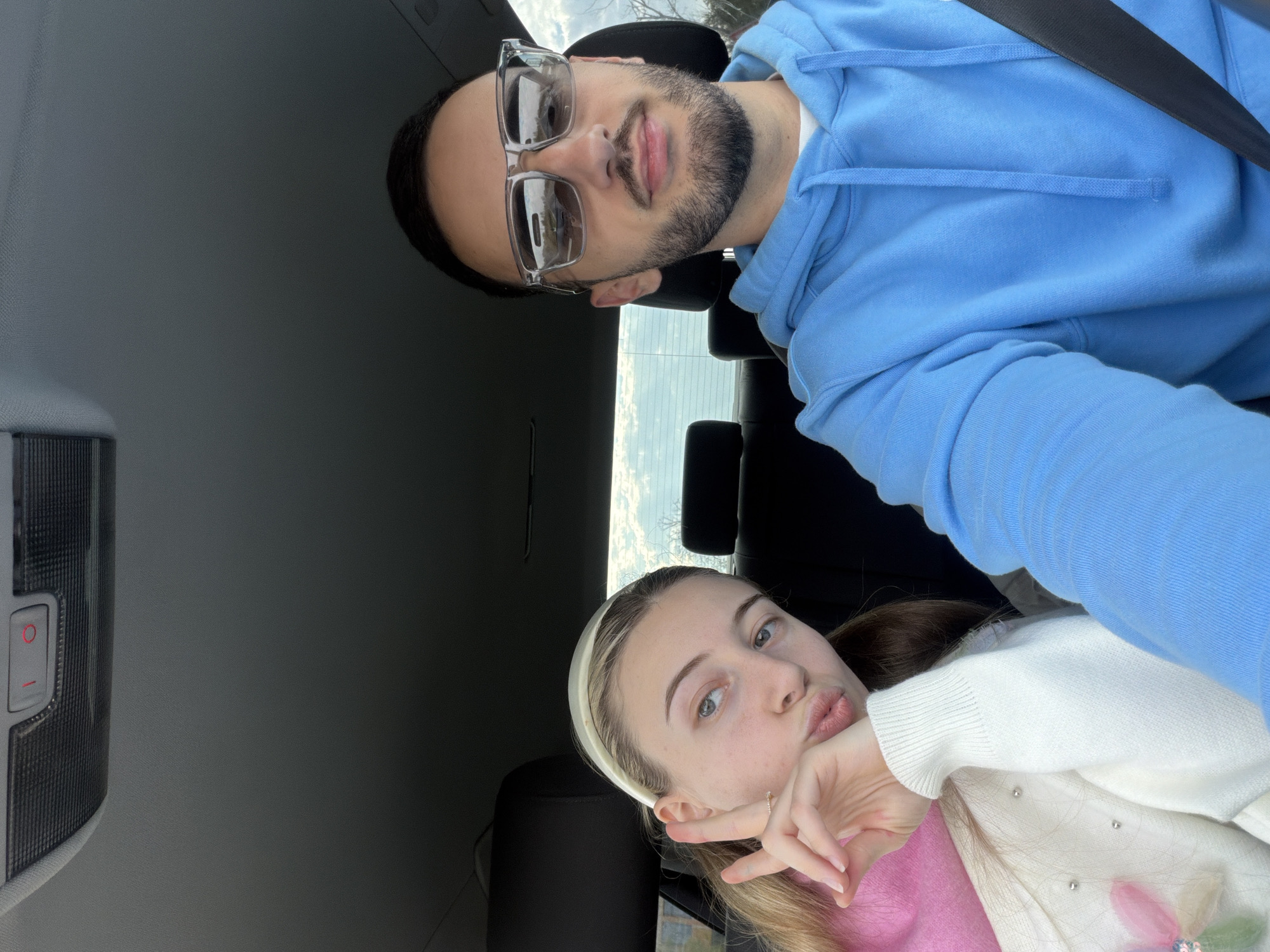 Two people are posing for a selfie inside a car. A man wearing a light blue hoodie and glasses is in the top portion of the frame, with a seatbelt visible across his chest. A woman with a white headband is in the lower portion of the frame, making a "peace" sign with her fingers and a kiss face. The car's interior is visible, including the ceiling and headrests. The car appears to be stationary, and judging by the scenery outside the window, it is daytime. It is not possible to determine the location within Plovdiv, Bulgaria from the information provided.