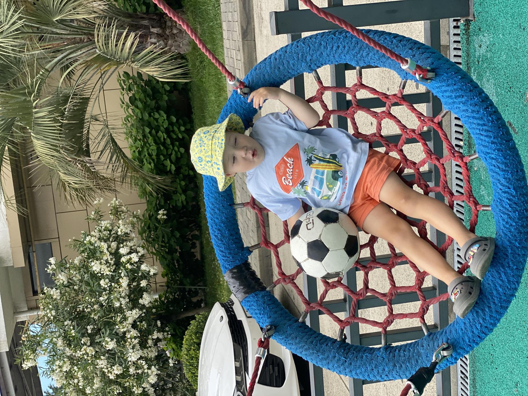 In Sochi, Russia, a young child wearing a yellow bucket hat with a marine motif and a light blue t-shirt with a beach scene graphic sits in a large, circular blue swing with a red net. The child is holding a black and white soccer ball. The child is wearing orange shorts and grey clogs with cartoon characters. The background shows lush green foliage, a white car, and part of a building. The swing is suspended by thick red ropes.