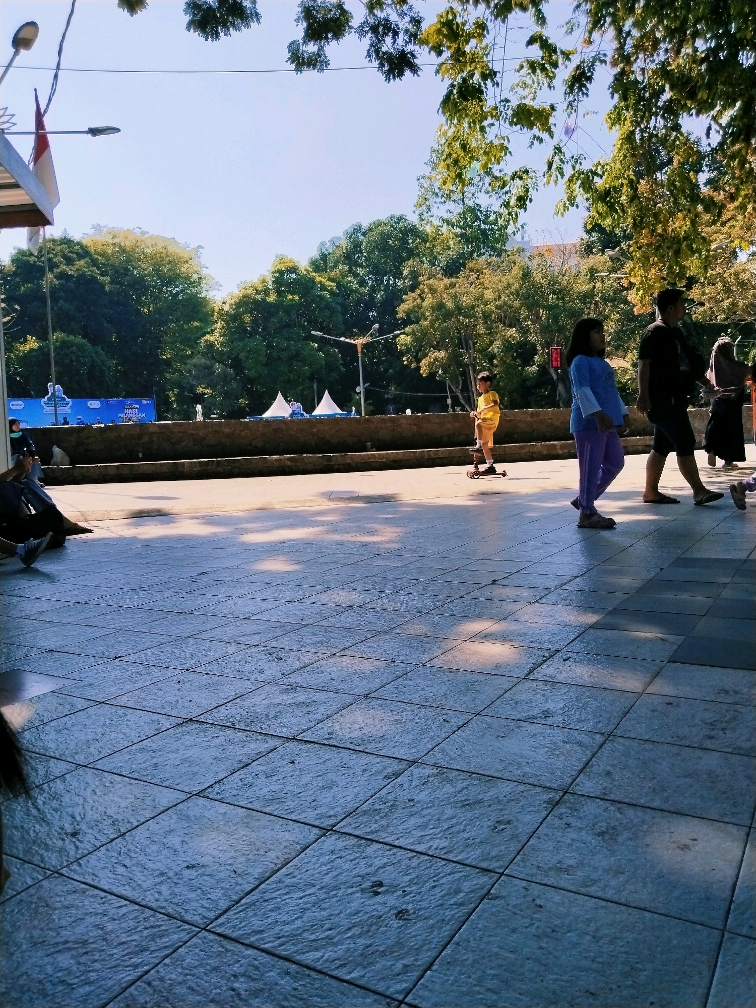 This image captures a scene in Surabaya, Indonesia, likely in a public park or plaza. The foreground is dominated by a tiled walkway, wet in places, with shadows of trees cast upon it, indicating the presence of sunlight filtering through foliage. In the midground, a group of people are visible. A young boy on a scooter, wearing a yellow shirt, is moving from left to right. Beside him, a girl in a blue shirt and purple pants is walking. Further to the right, a man in a dark t-shirt and shorts is walking, followed by a woman in a hijab. In the background, there is a stone wall or barrier, beyond which are more trees and some structures, possibly tents or booths, set up for an event. A blue banner with text, possibly advertising, is also visible. The overall atmosphere suggests a sunny day in a lively public space.