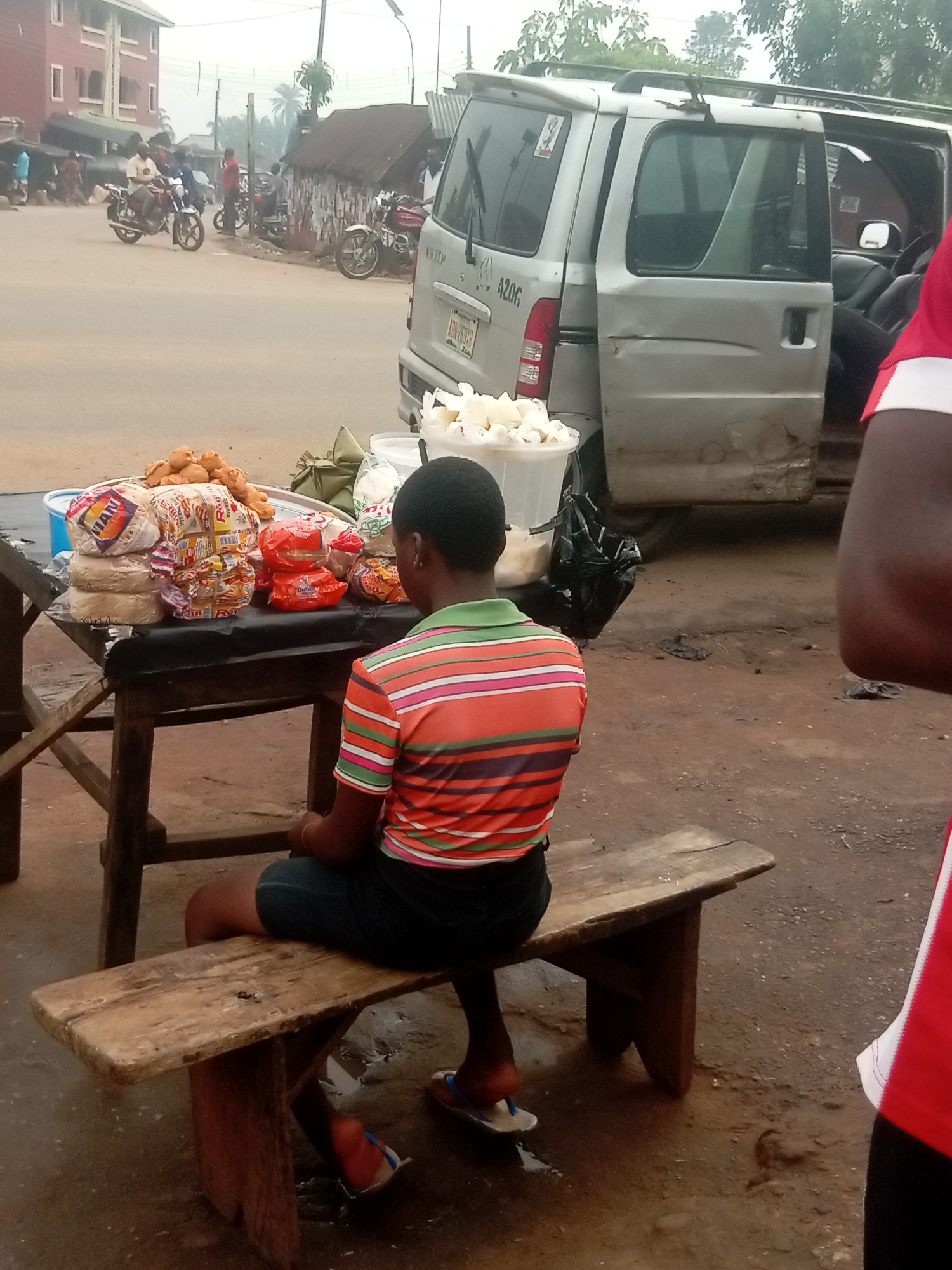 A street scene in Nkpor, Nigeria, shows a young person sitting on a wooden bench at a table laden with bread, pastries, and other food items for sale. The vendor is wearing a striped shirt and dark shorts. Behind them is a damaged silver van with its door open, and another person partially visible in a red shirt. In the background, there's a street with motorcycles and buildings. The ground is dirty and wet.