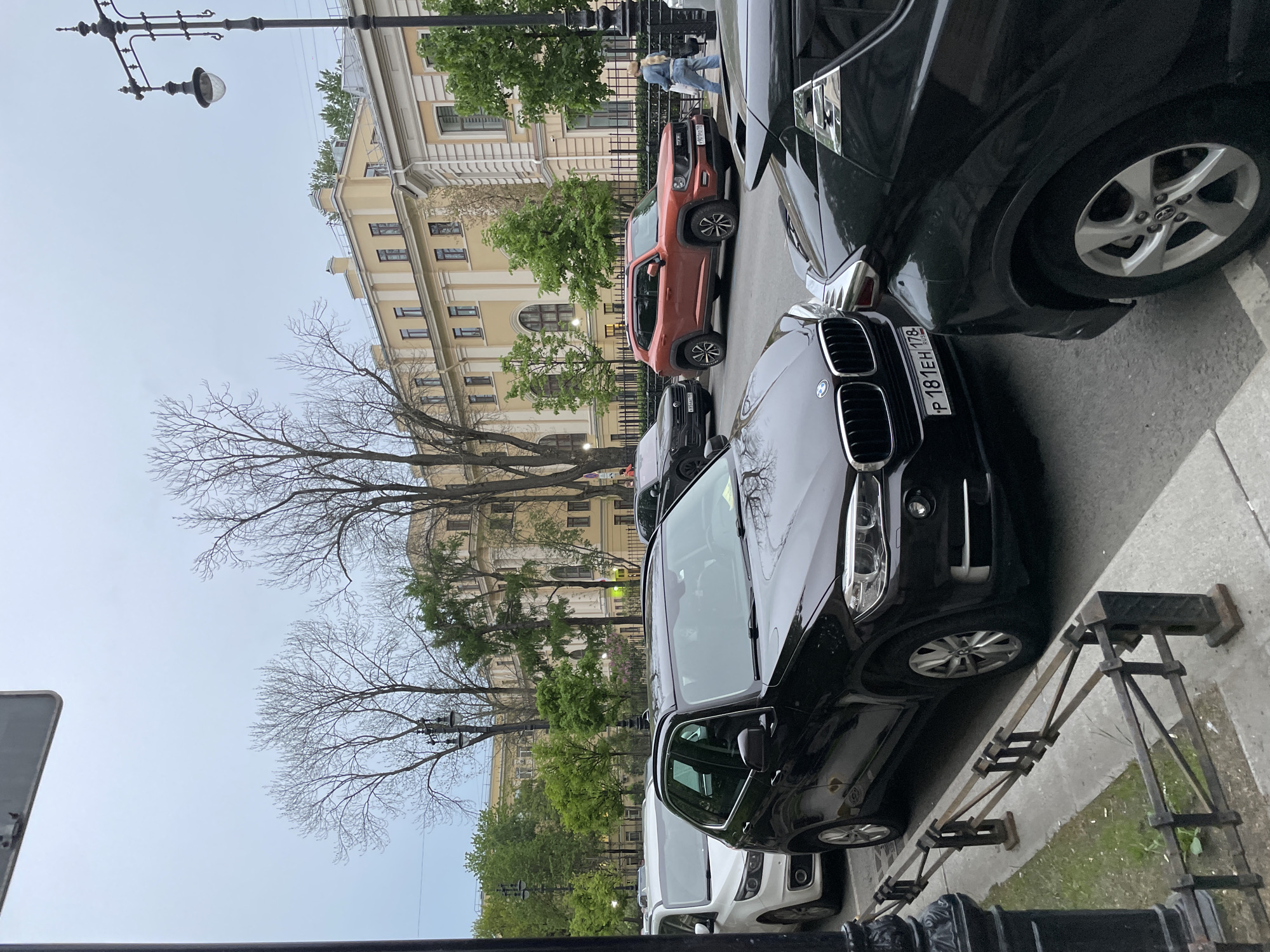 The image shows a street in Saint Petersburg, Russia, lined with parked cars. The cars are a mix of colors including black, white, and orange. A light post is on the left, and a building with windows is visible in the background. Some trees with bare branches are also visible. A person can be seen in the distance.