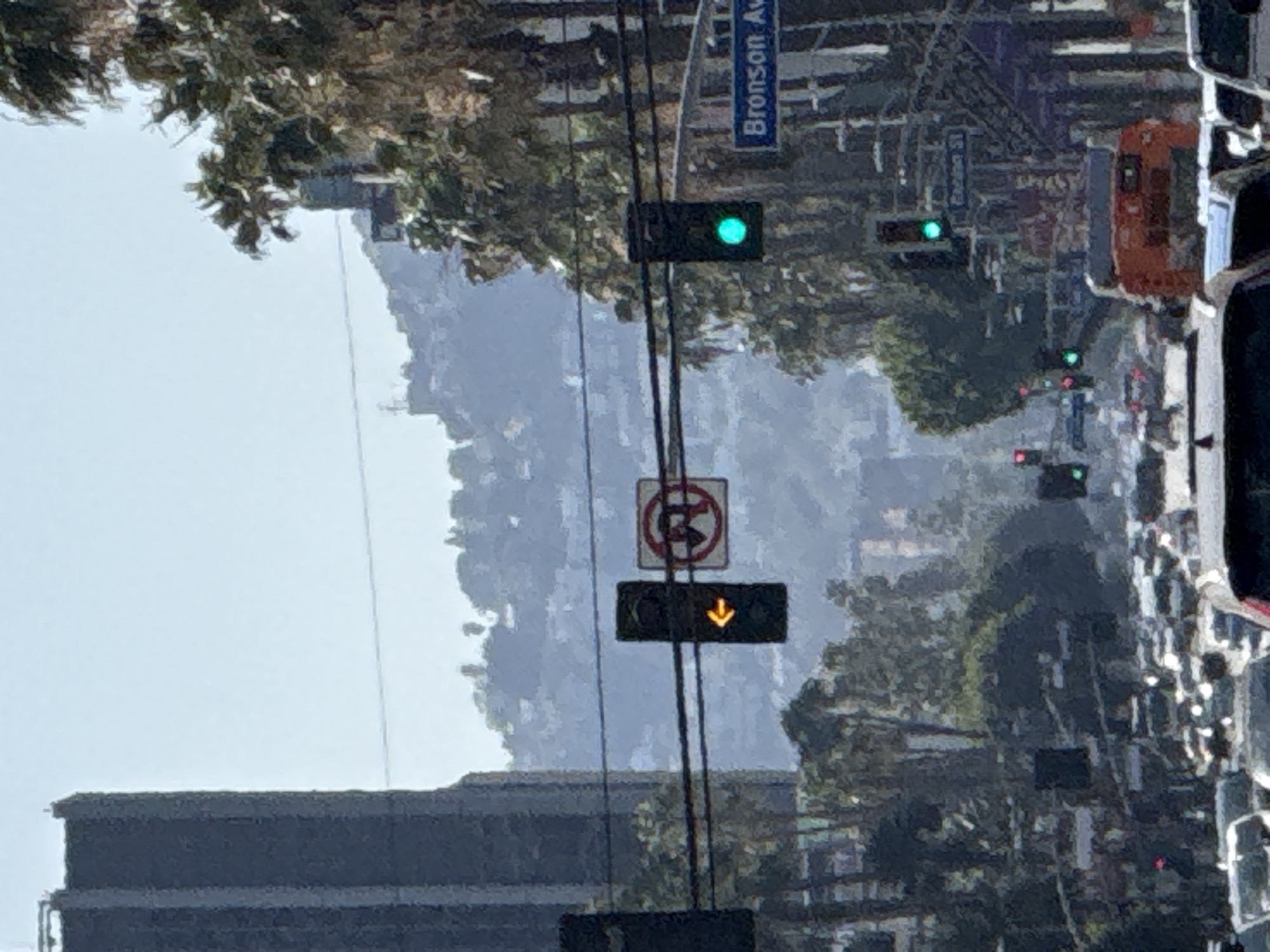 The image shows a street scene in Los Angeles, California, likely on Bronson Avenue due to the visible street sign. There's traffic on the right side of the frame. A traffic signal displays a green light, with other signals further down the road showing green and red. A yellow arrow light is present on a utility pole with a no parking sign above it. Hills are visible in the background. A building is in the foreground on the left side of the frame. Utility poles and lines cross the scene.